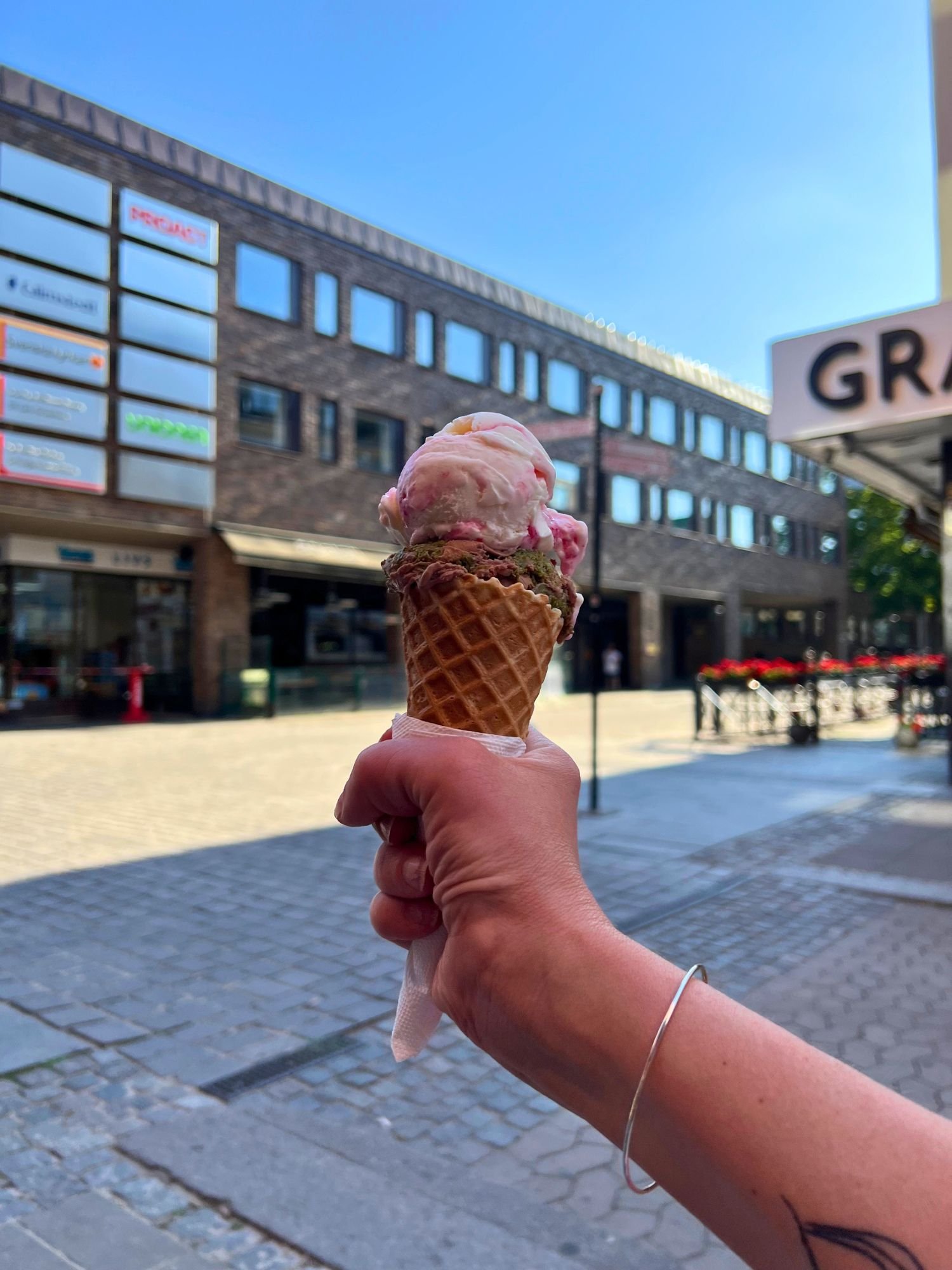 A hand holding a waffle cone with two scoops of ice cream, chocolate-pistachio Dubai chocolate and pink and white polka, set against a sunny, blurred city street background.