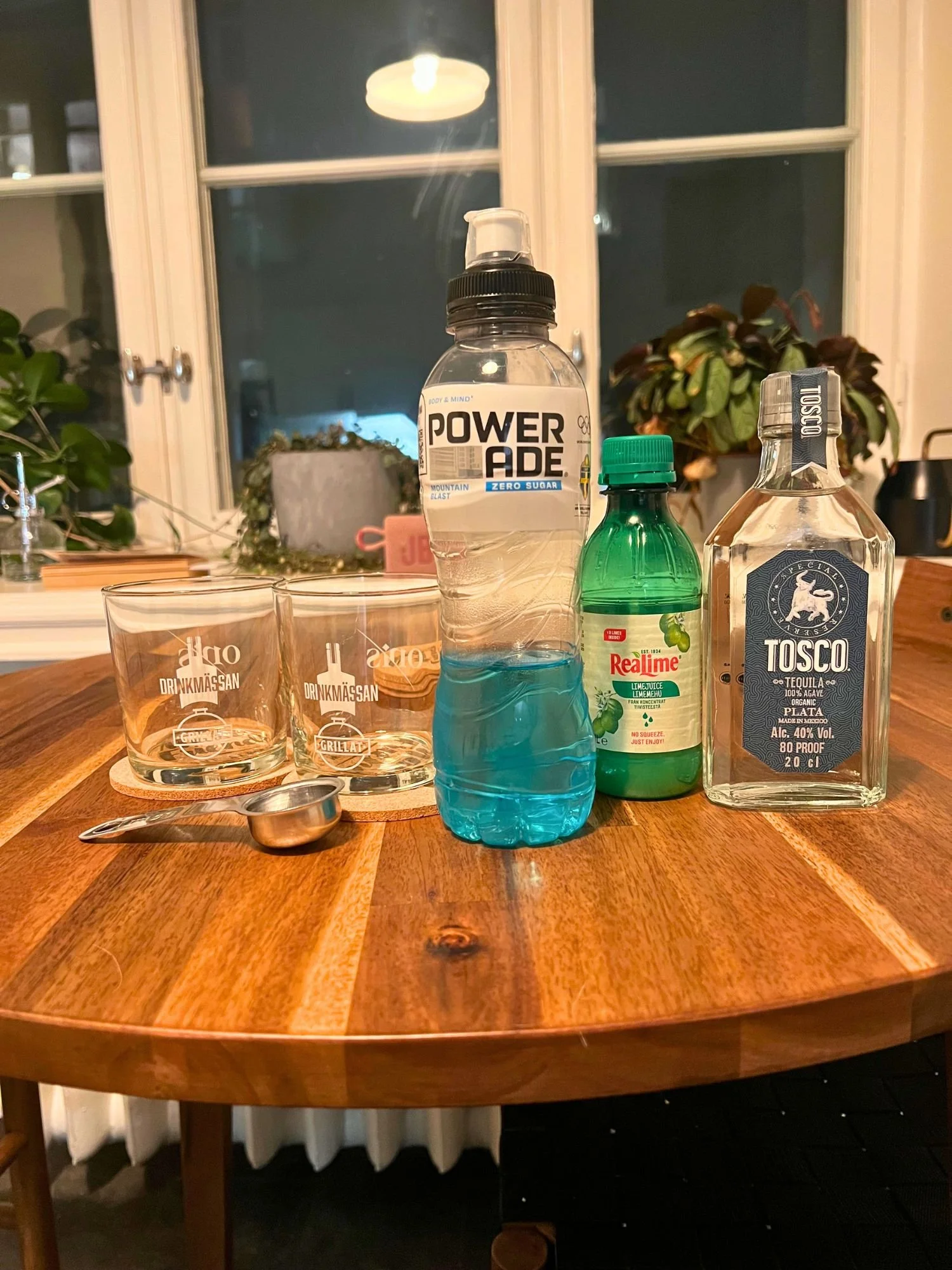 Ingredients for a blue cocktail on a wooden table, including blue Powerade, tequila, lime juice, measuring tools, and glasses, with a window and plants in the background.