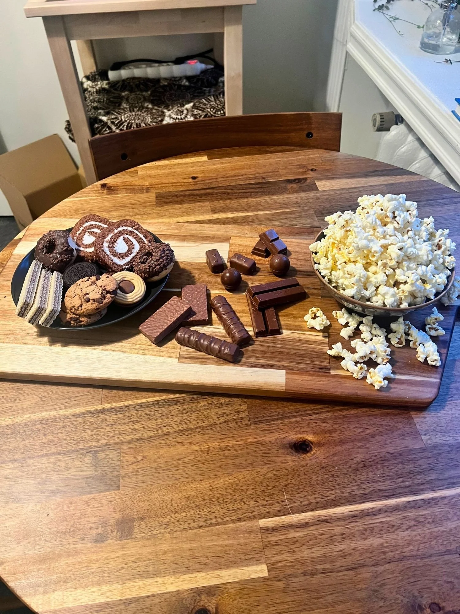 High-angle view of a wooden serving board on a round wooden table, arranged with a bowl of popcorn, assorted chocolates, and a plate of cookies and mini donuts.