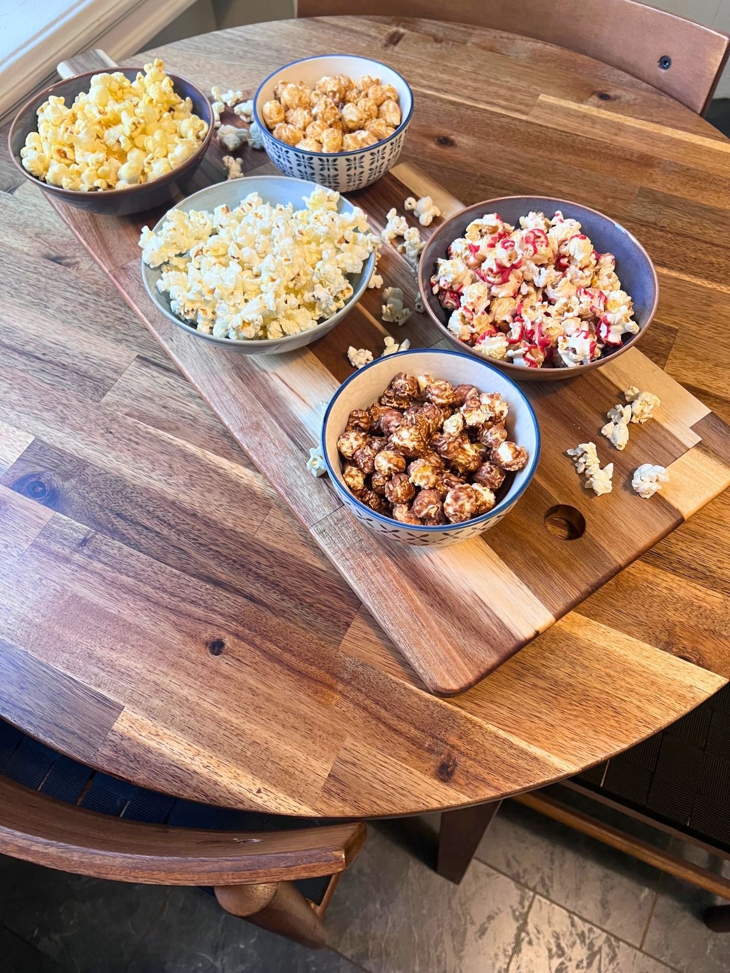 High-angle shot of five ceramic bowls filled with different varieties of gourmet popcorn set on a wooden serving board across a round wooden table.