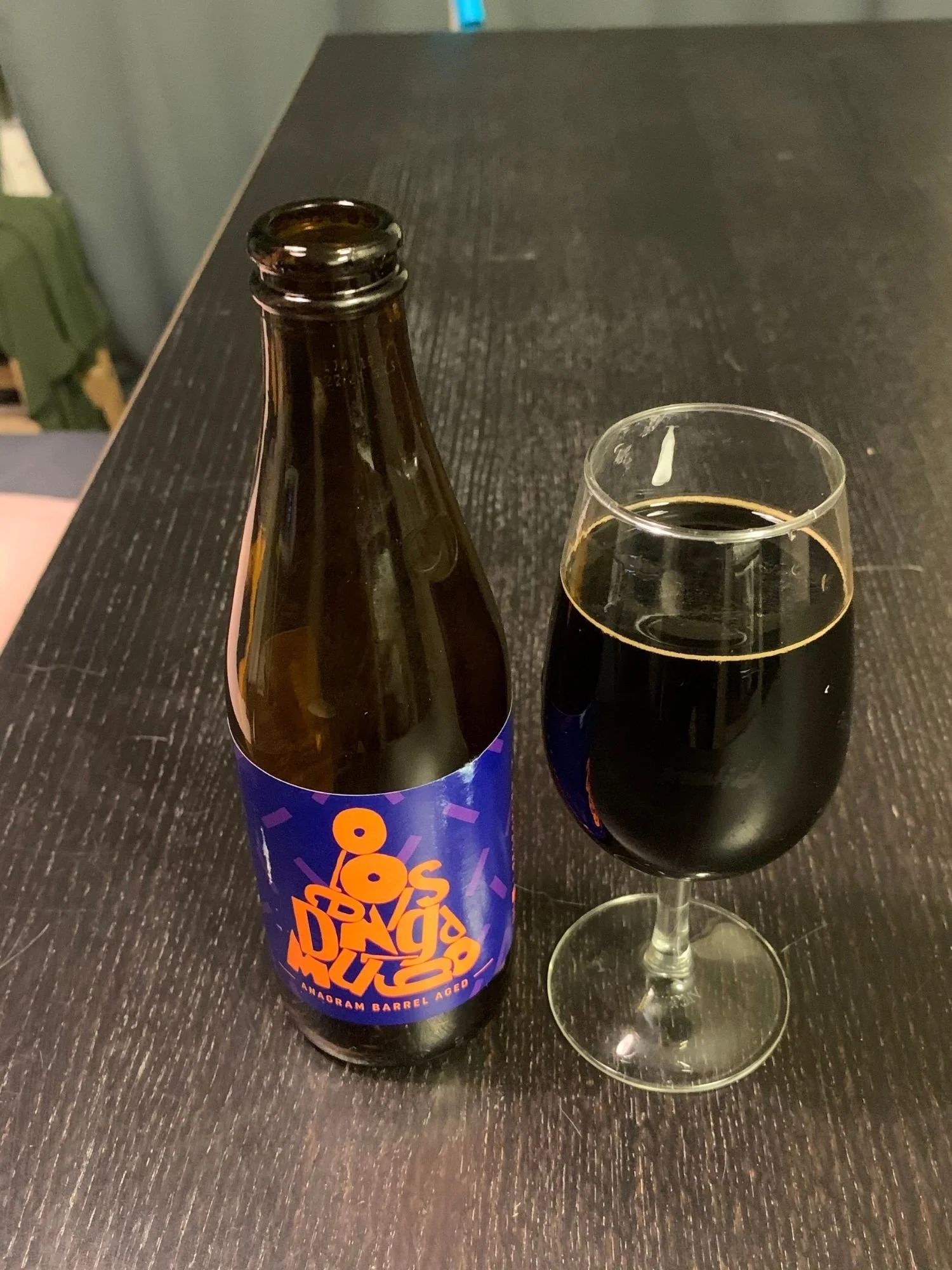 A brown glass beer bottle next to a tulip glass filled with dark imperial stout on a dark wooden tabletop.