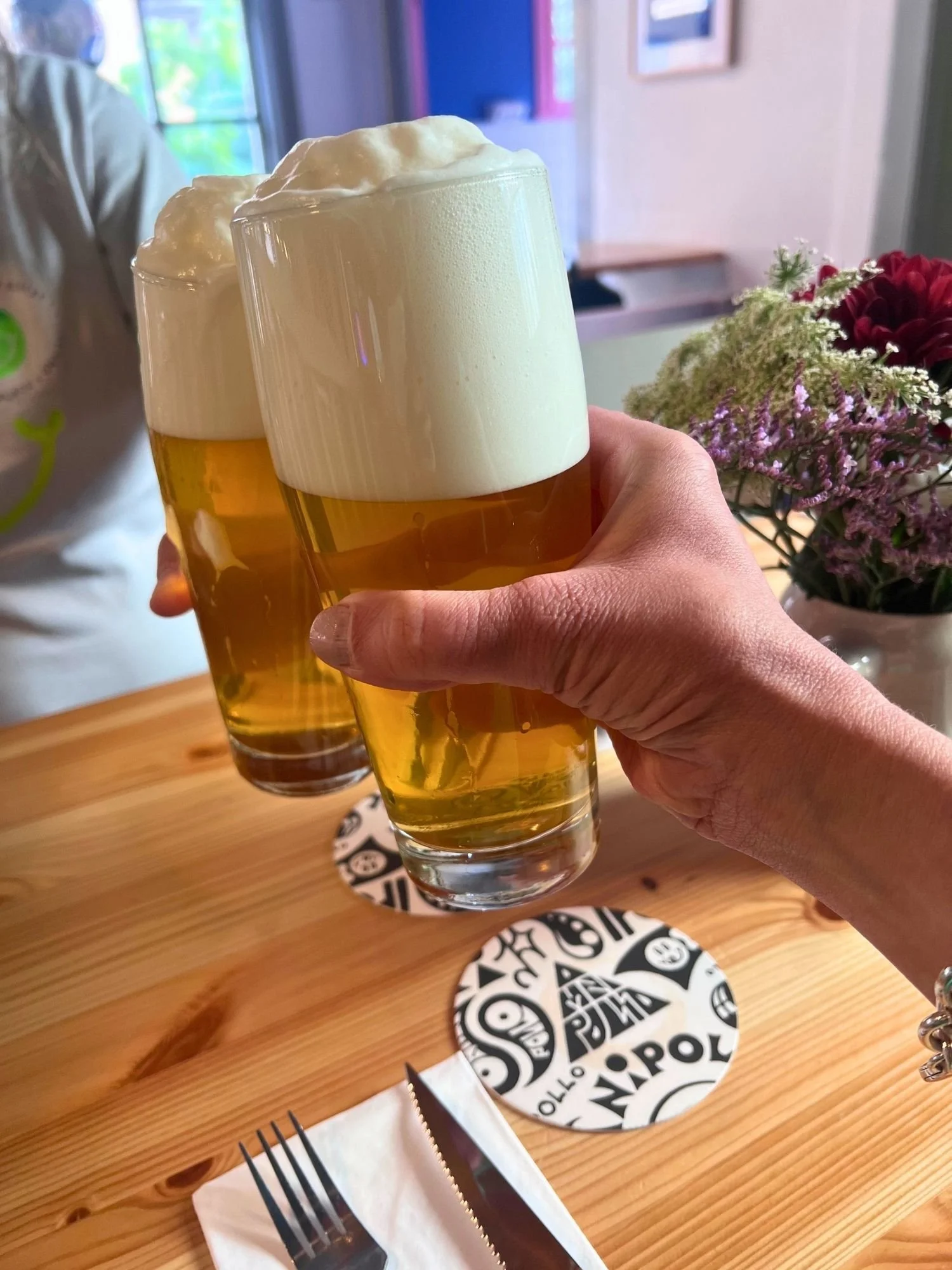 A close-up view of two hands clinking glasses of golden draft beer together in a toast over a light wooden table. Both glasses are topped with thick foam crowns.
