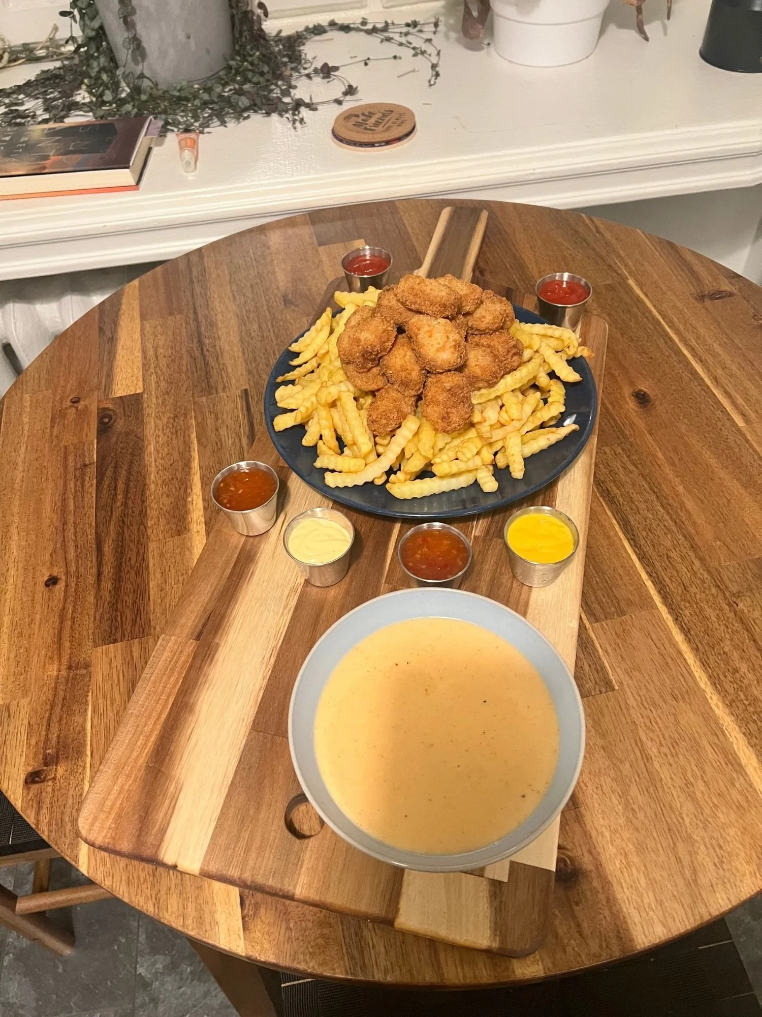 A wooden serving board holds crinkle-cut fries topped with golden vegetarian nuggets, accompanied by small cups of ketchup, mustard, and other dipping sauces, with a bowl of creamy cheese dip beside it.