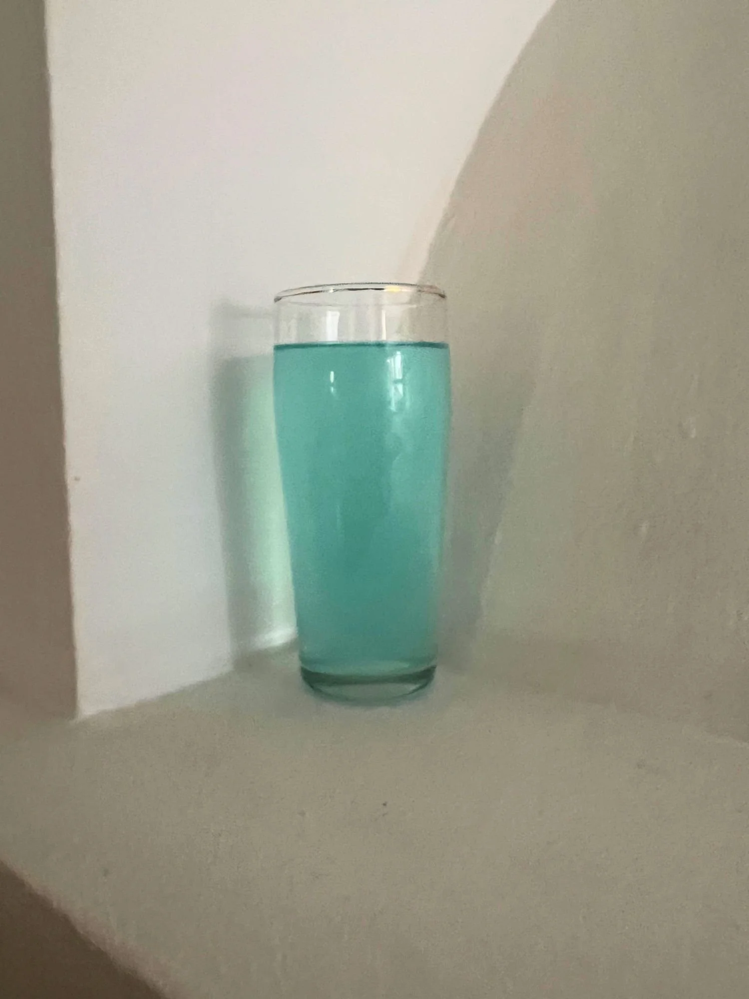 Tall glass filled with a light blue cocktail placed in a corner against a white wall.