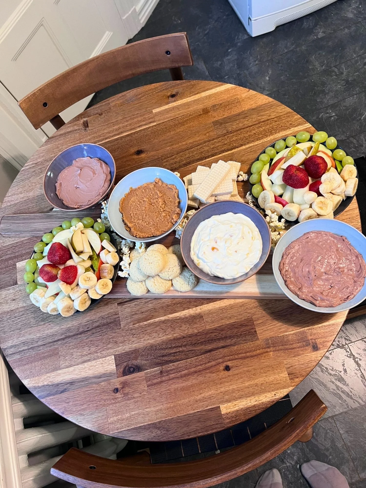 High-angle view of a wooden serving board set across a round wooden table, laden with bowls of sweet dessert dips, fresh fruit platters, cookies, and vanilla wafers.