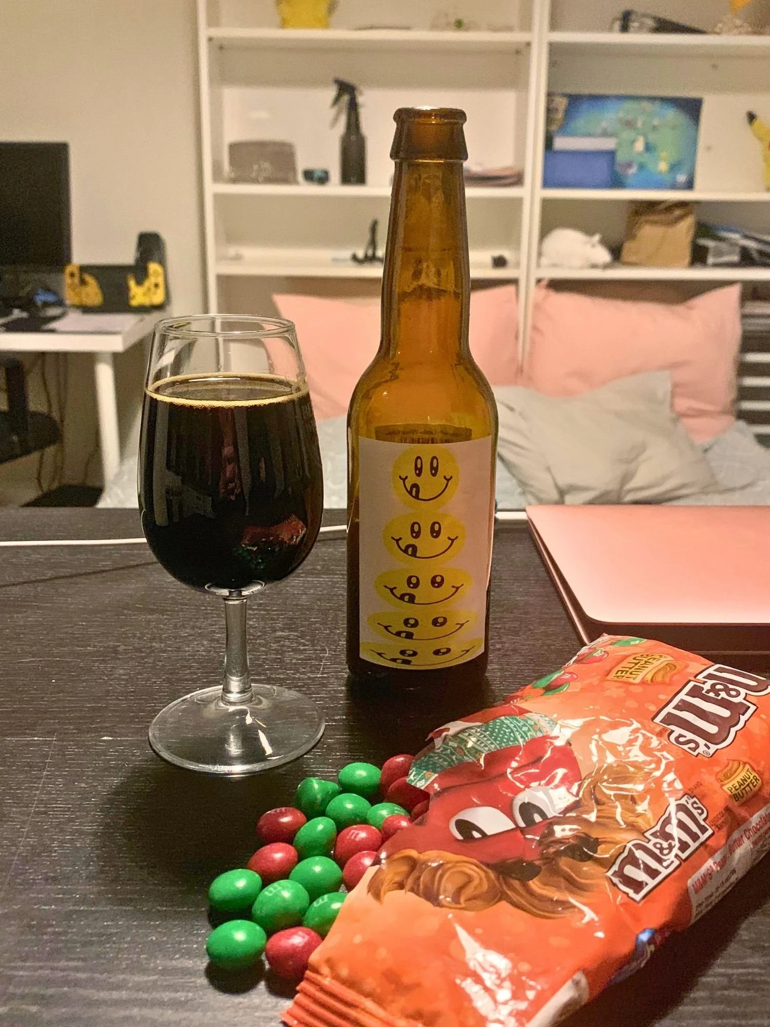 A brown beer bottle with a white label with a vertical stack of yellow smiley faces next to a glass filled with dark stout on a dark table.