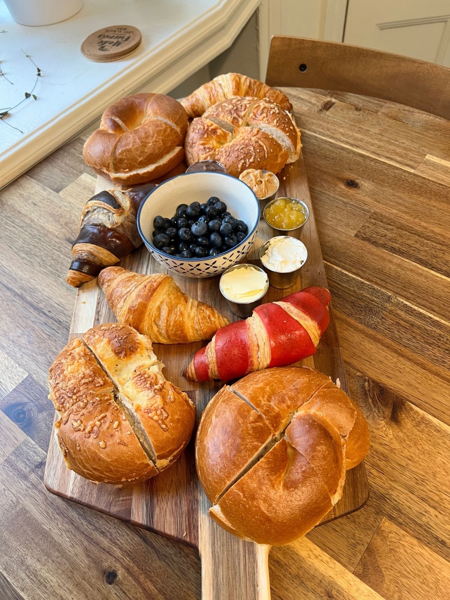 A long wooden serving board rests vertically on a round wooden table, presenting a breakfast board filled with croissants, bagels, and spreads.
