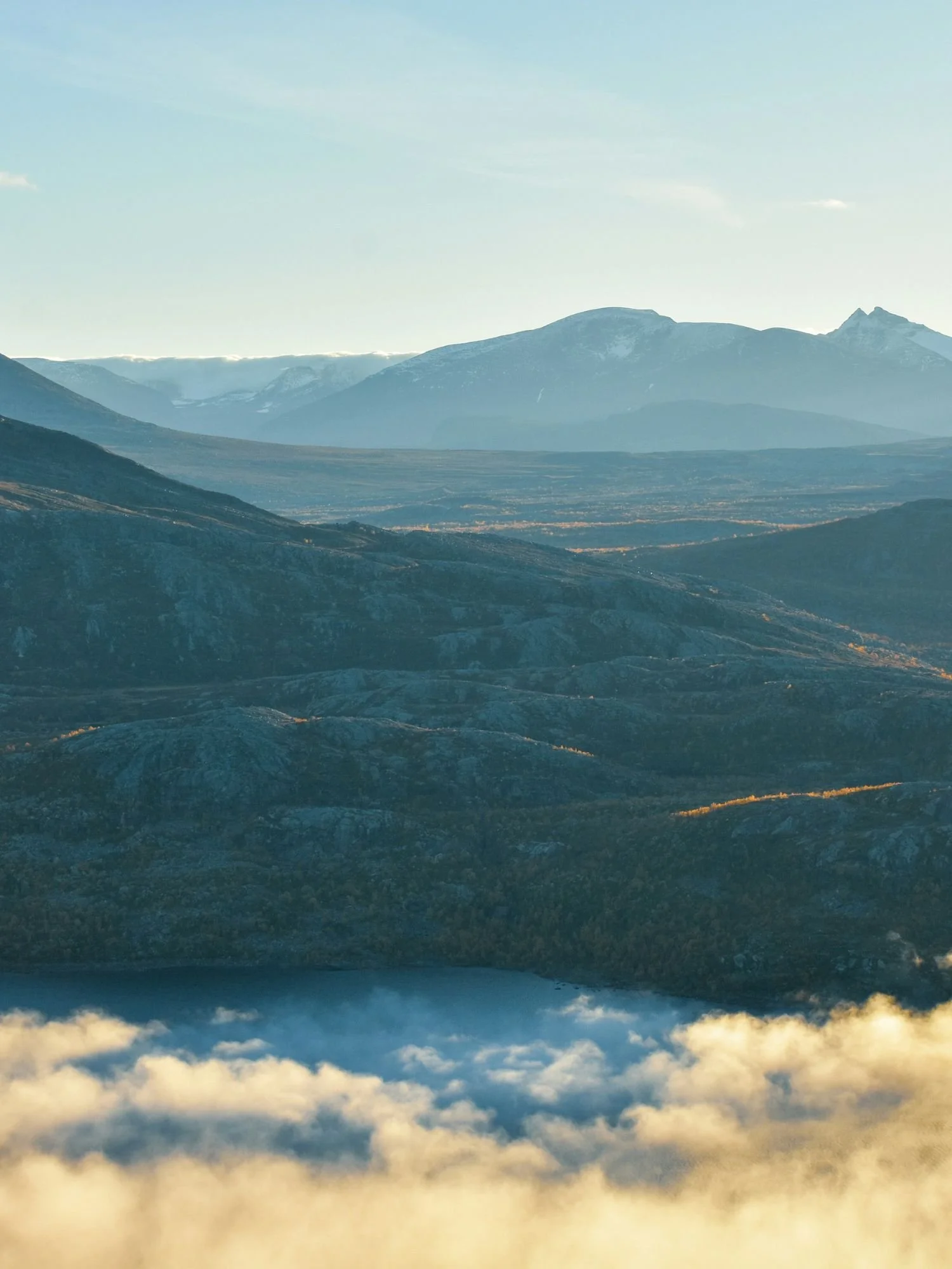 A high-angle, sweeping landscape of Stora Sjöfallet National Park showing rolling, rocky mountain ridges above a low layer of clouds illuminated by golden sunlight.
