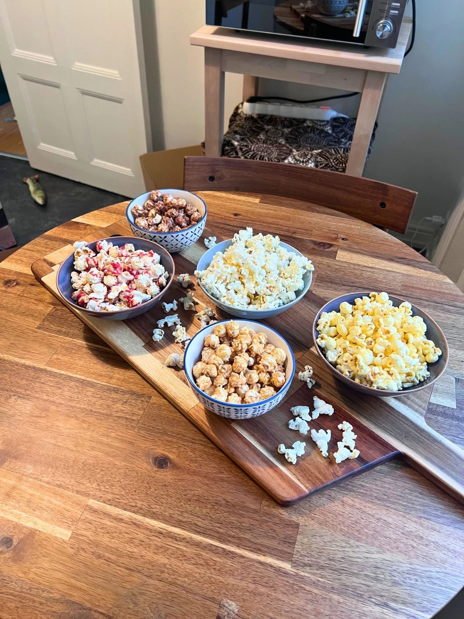 A wooden table set with a long serving board containing five bowls of different flavored popcorns, including caramel, chocolate, peanut butter and jelly, cheese, and plain.