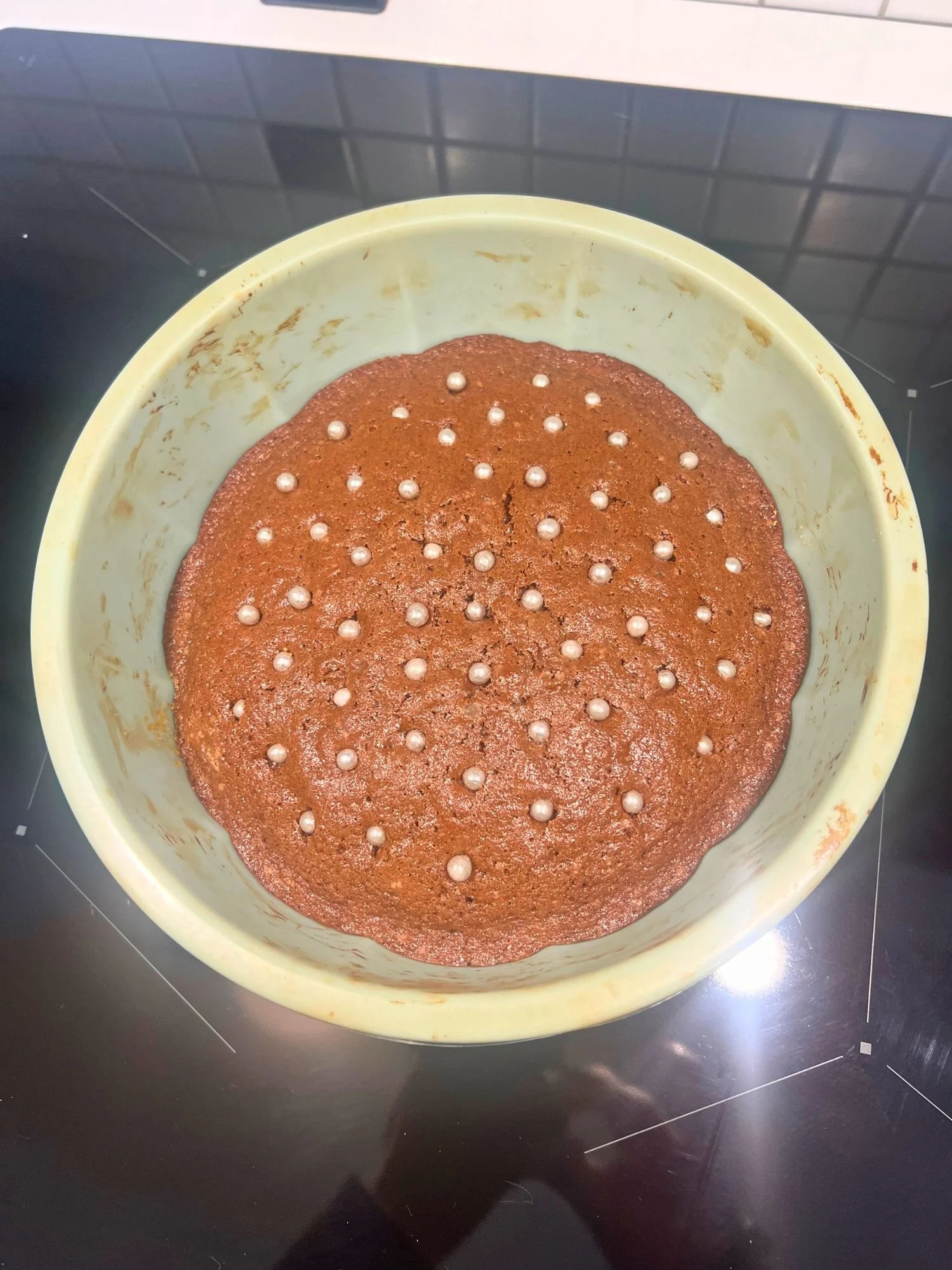 Freshly baked chocolate sockerkaka in a light green round baking pan, topped with evenly scattered round, silver cake decorations, sitting on a black stovetop.