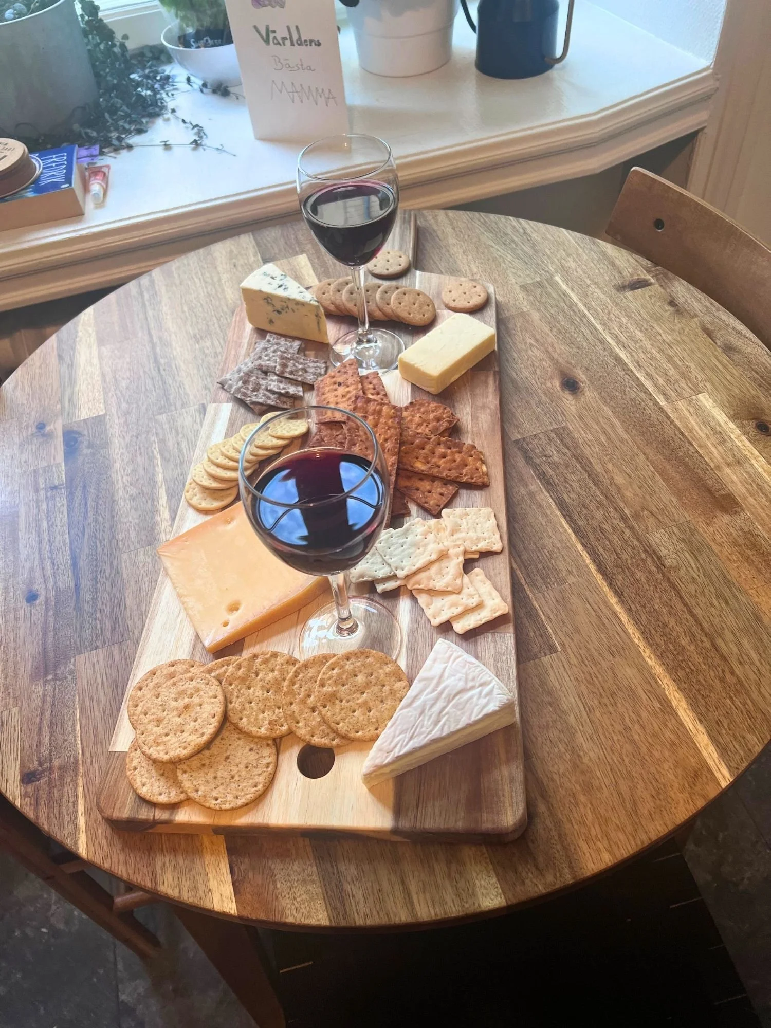 High-angle view of a long wooden board filled with various cheeses, crackers, and two glasses of red wine on a round wooden table.