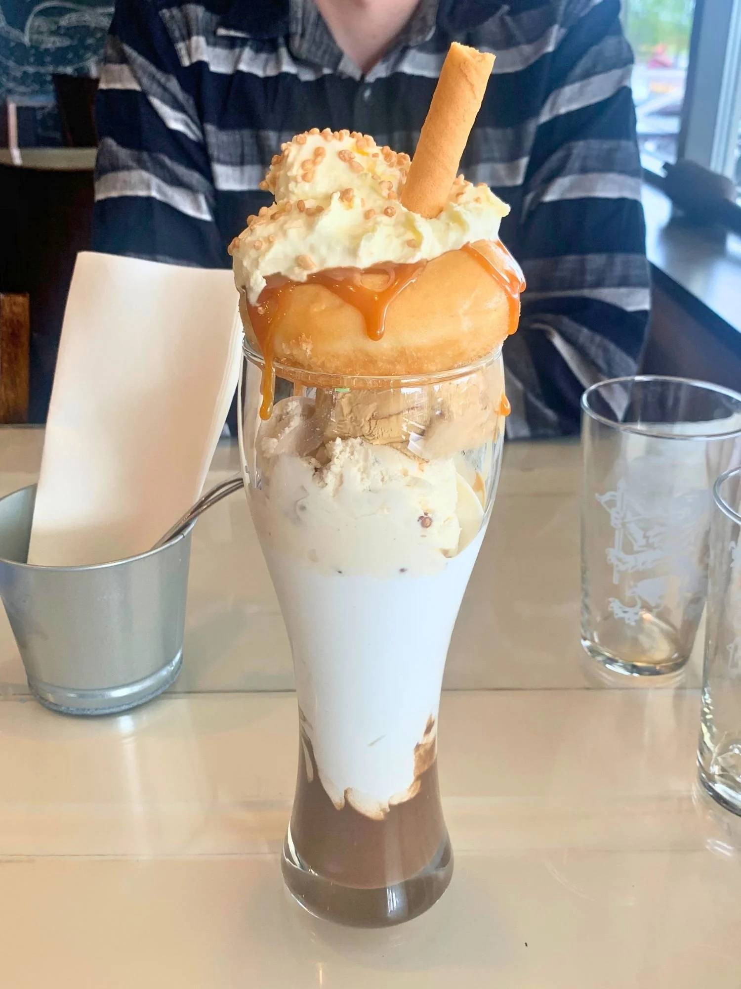 An over-the-top ice cream milkshake in a tall glass, capped with a whole glazed donut, whipped cream, caramel sauce, and a wafer stick.
