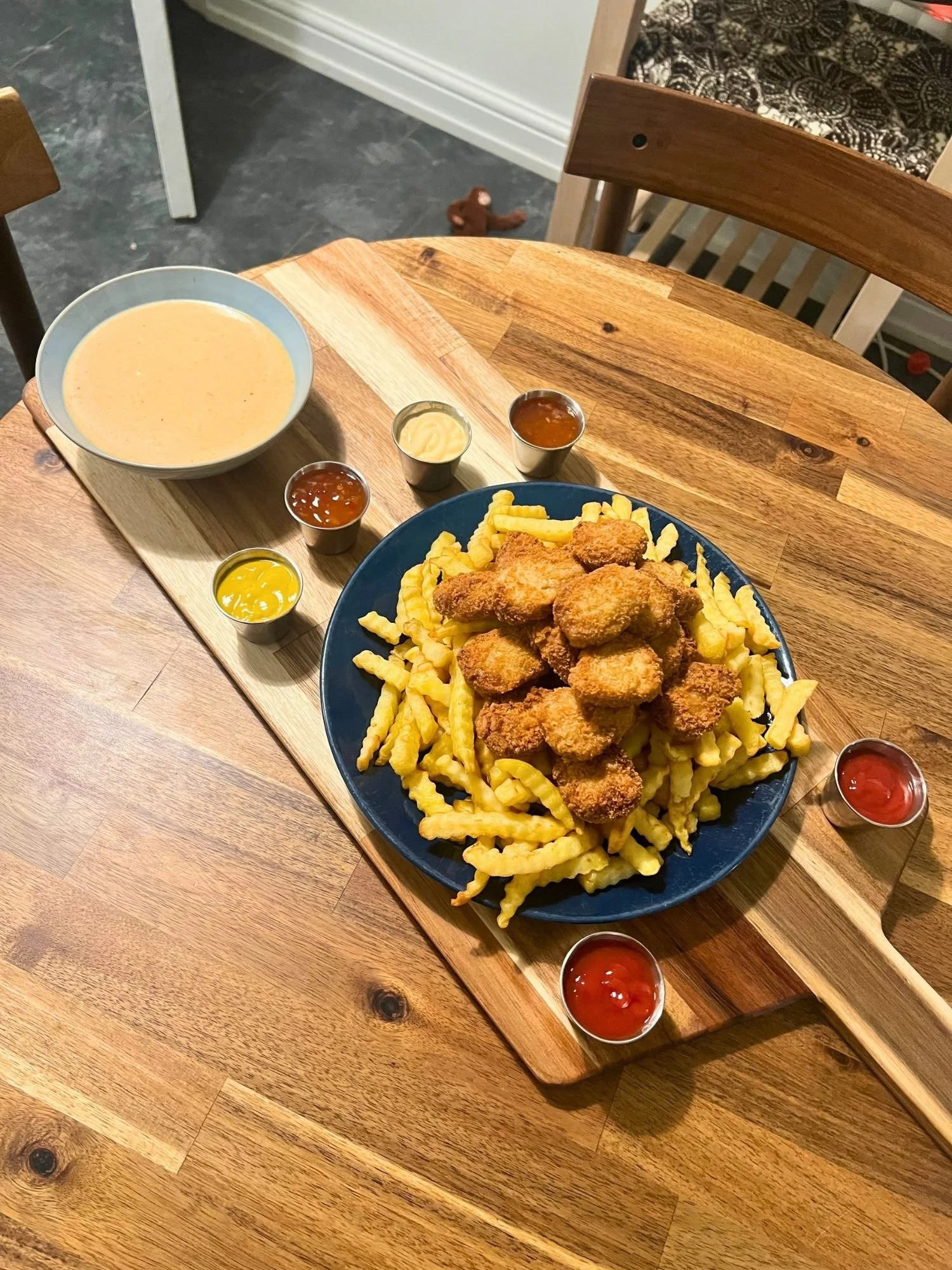 A wooden serving board on a round wooden table holding a large blue plate stacked with crinkle-cut fries and nuggets, surrounded by a bowl of cheese dip and various dipping sauces.