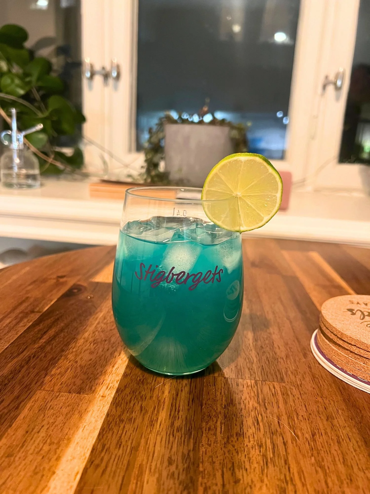 Glass of blue cocktail over ice garnished with a lime slice, sitting on a wooden table with a window and houseplants in the background.