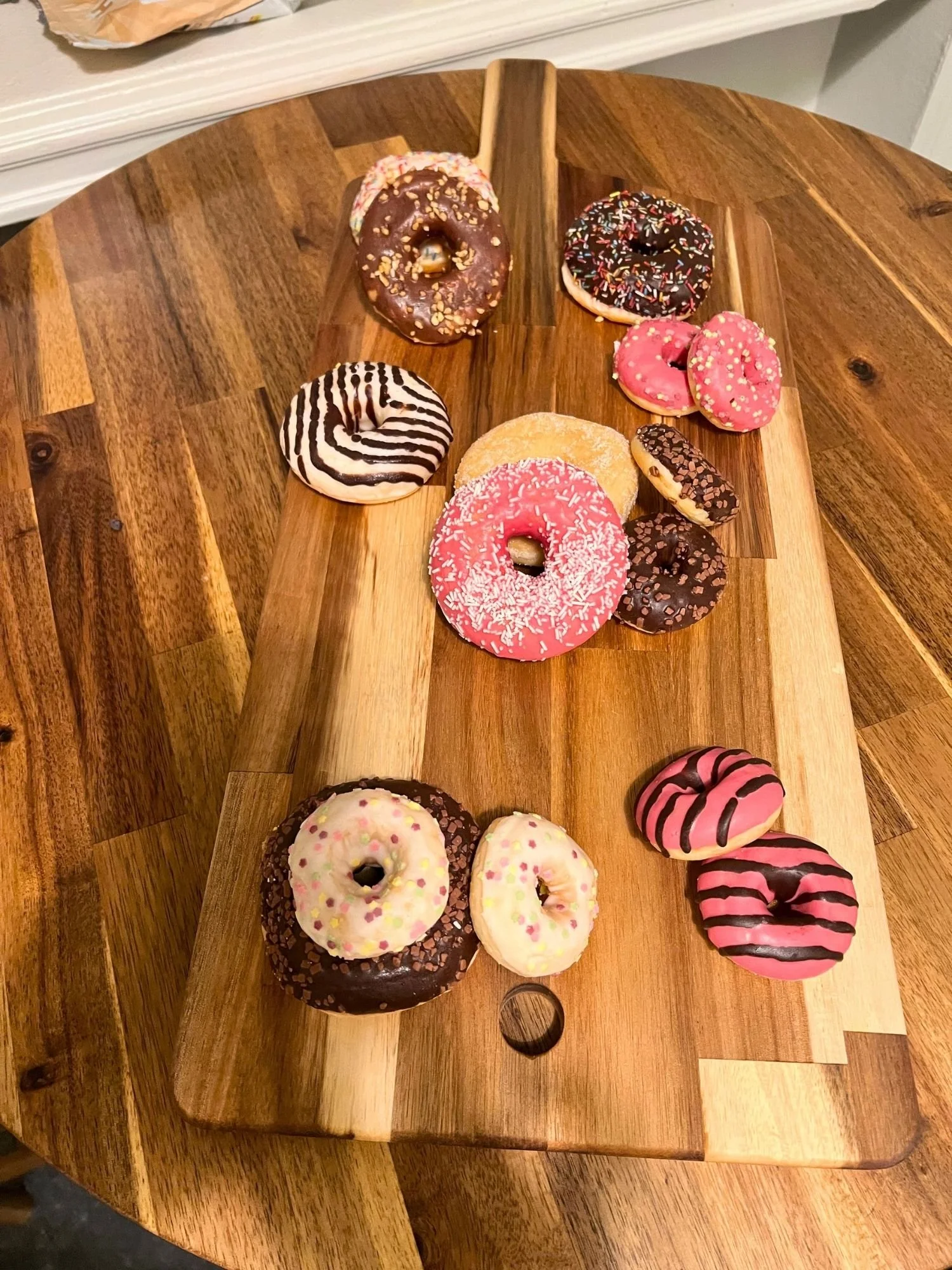 A long wooden serving board rests on a round wooden table, arranged with a variety of colorful glazed and frosted donuts.