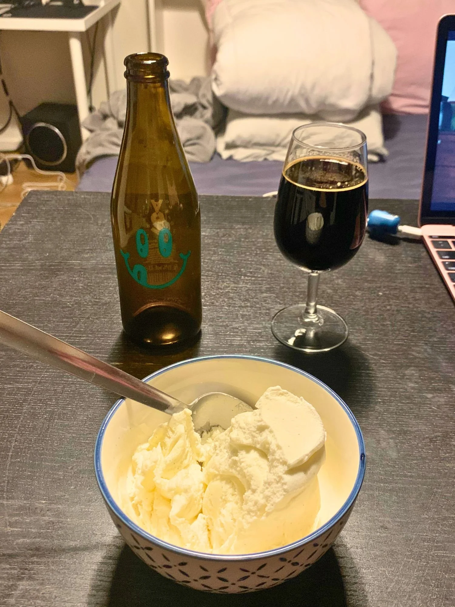 A bowl of vanilla ice cream with a spoon on a dark wooden table next to an open brown glass bottle with a teal smiley face logo and a stemmed glass filled with a dark imperial stout.