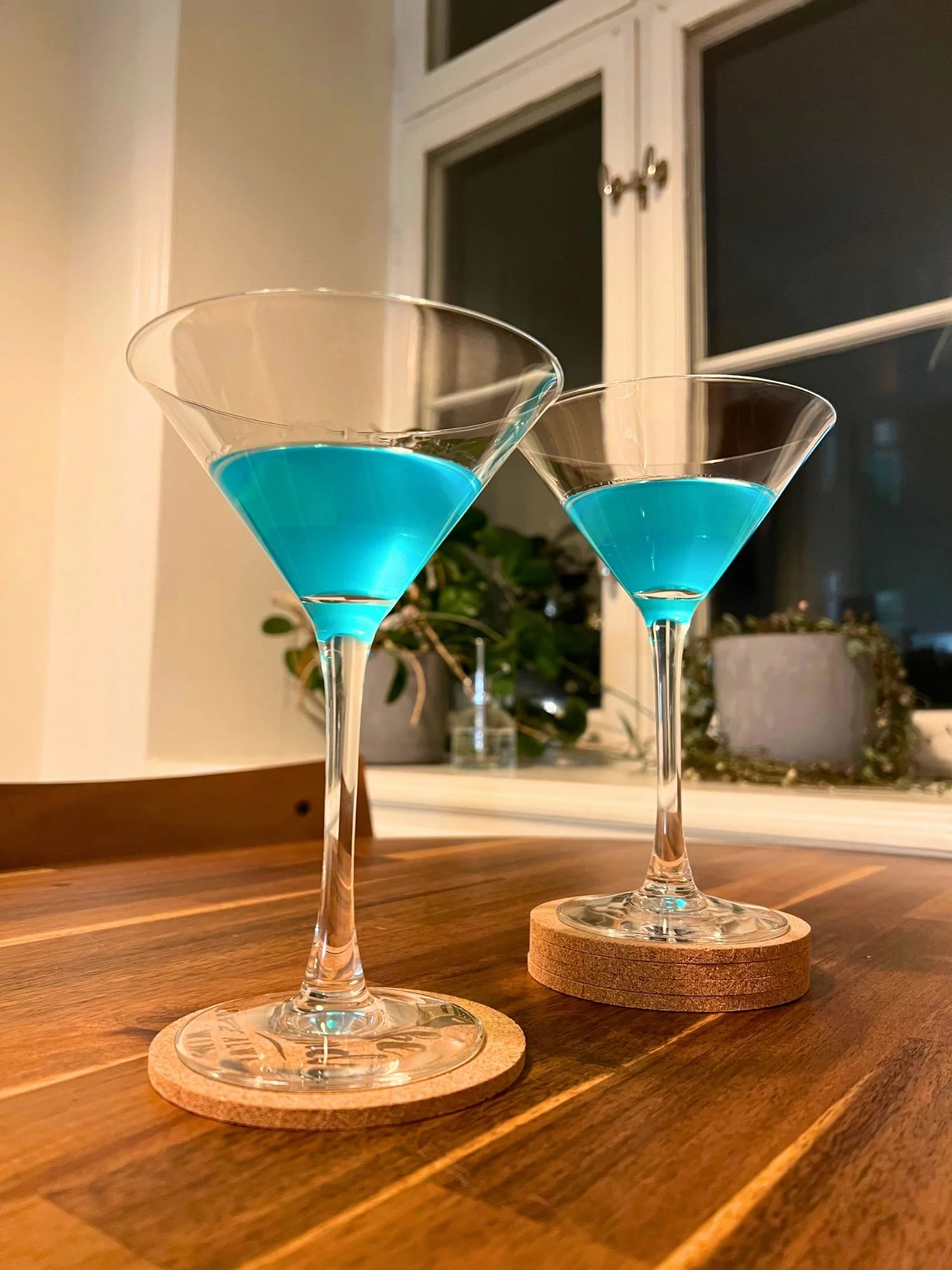 Two martini glasses filled with bright blue cocktails, placed on a wooden table with a window and plants in the background.