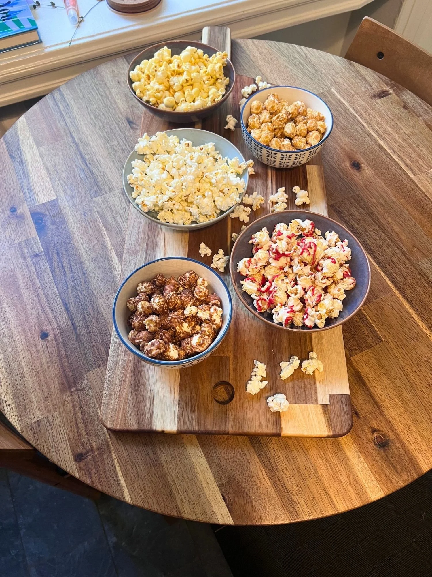 High-angle, length-wise shot of five ceramic bowls containing different varieties of gourmet popcorn set on a wooden serving board on a round wooden table.