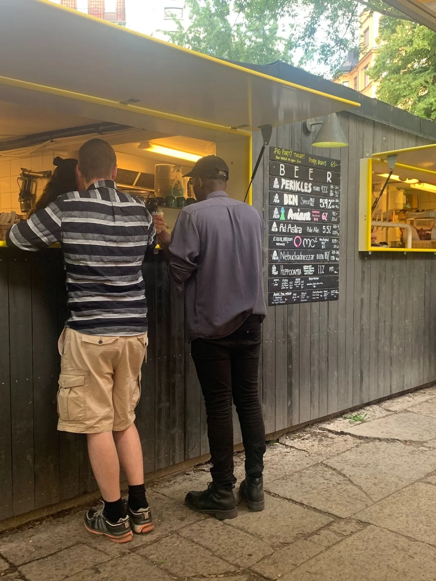 Two patrons stand facing an outdoor service counter built into a grey wooden food stand with yellow trim. To their right, a black chalkboard menu mounted on the exterior wall lists various beers with their style, ABV, and prices.