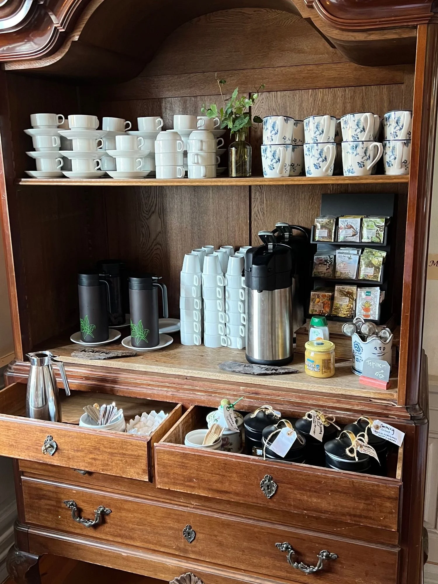 A self-service tea and coffee station in a vintage wooden hutch at afternoon tea at Vadstena Klosterhotel. The top shelf has neatly stacked white porcelain teacups and saucers on the left and white mugs with blue floral patterns are on the right.