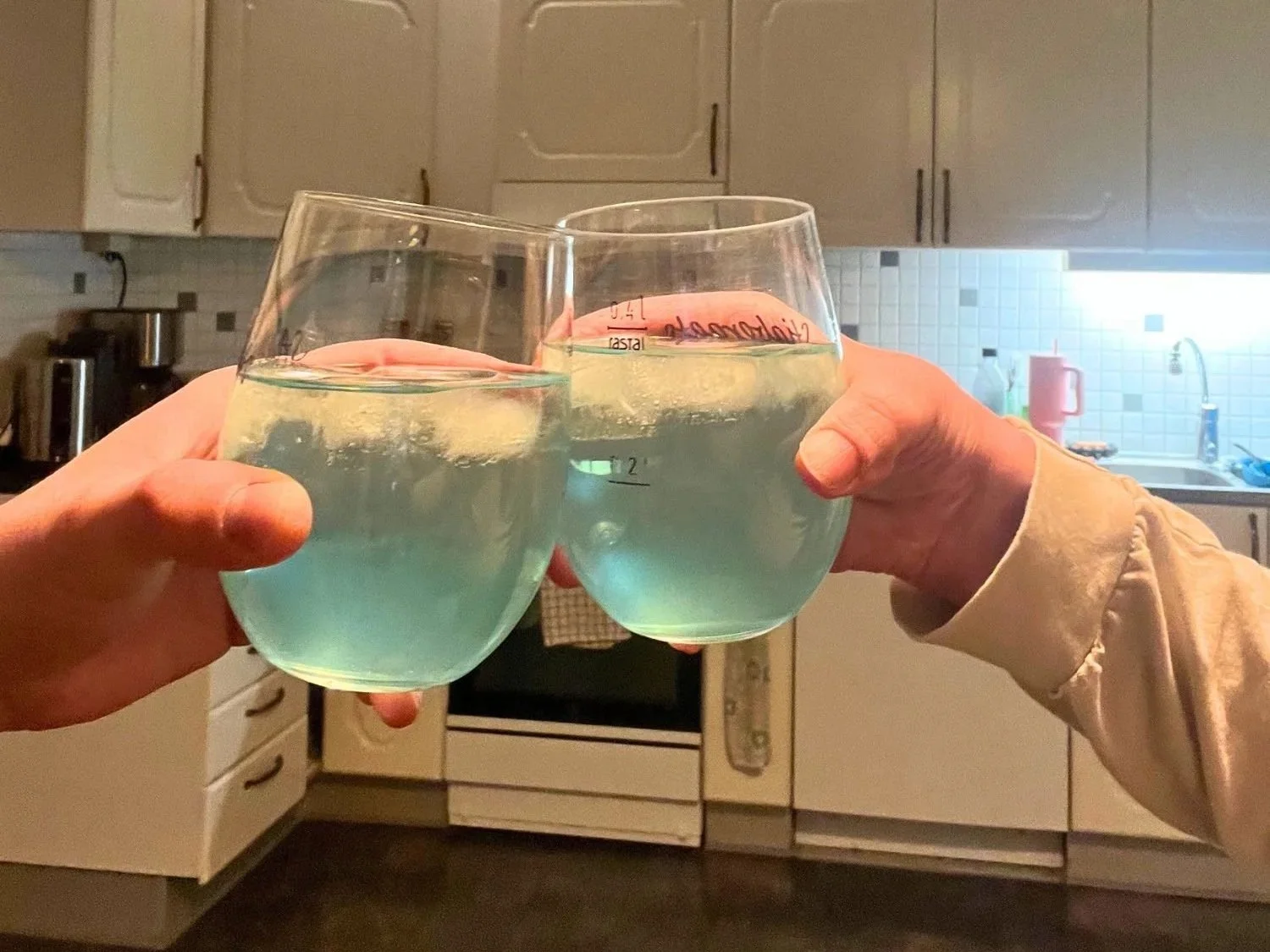 Two hands holding and clinking glasses of light blue cocktails with ice in a kitchen setting.