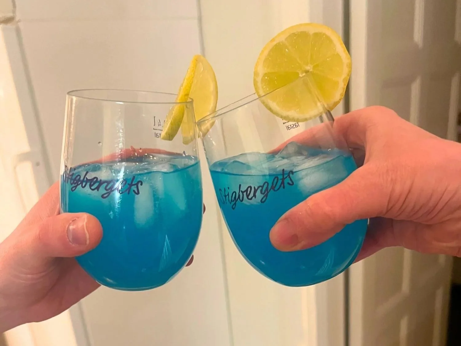 Two hands holding and clinking glasses of bright blue cocktails over ice, garnished with lemon slices, in a kitchen setting.