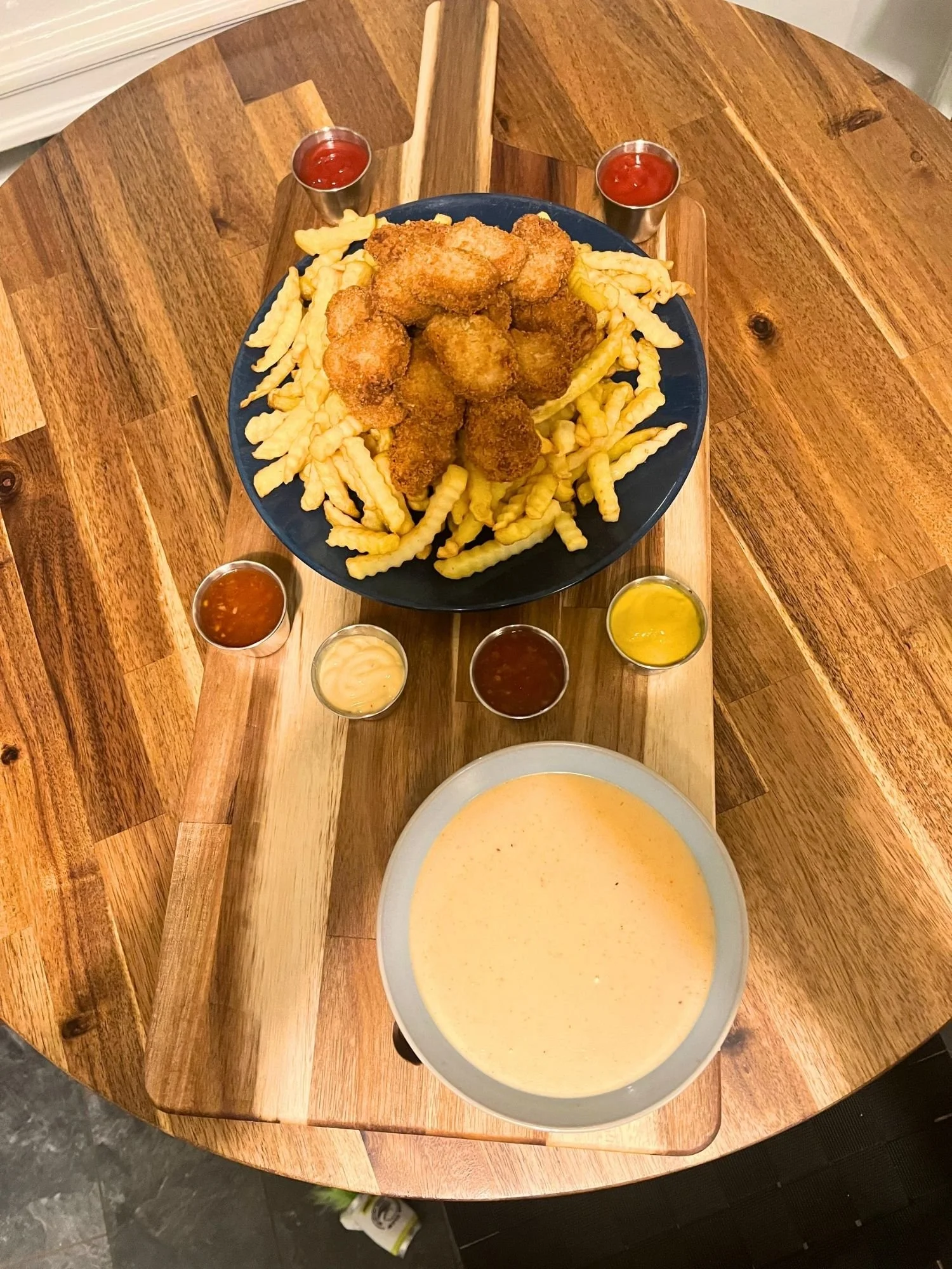 A long wooden serving board lies vertically across a round wooden table, presenting a large blue plate filled with golden crinkle-cut French fries and crispy fried nuggets piled on top.