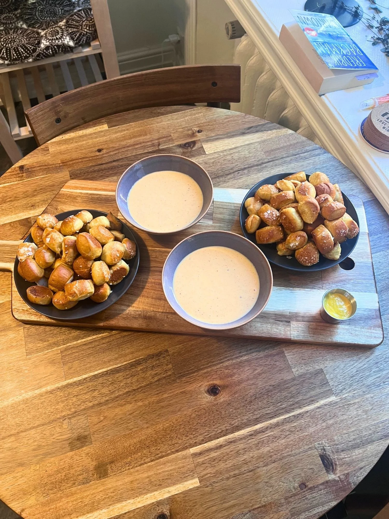 A wooden table set with a serving paddle holding two plates of warm pretzel bites, two bowls of cheese dip, and a small metal cup of yellow mustard.