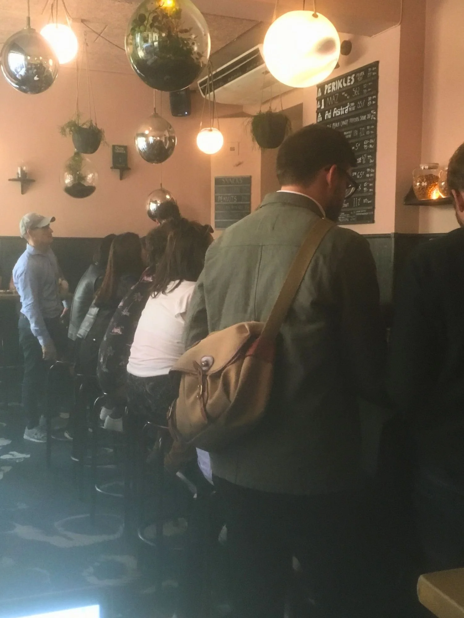 A view of patrons inside a dimly lit bar, seen from behind a man wearing an olive-green jacket and a tan crossbody canvas bag.