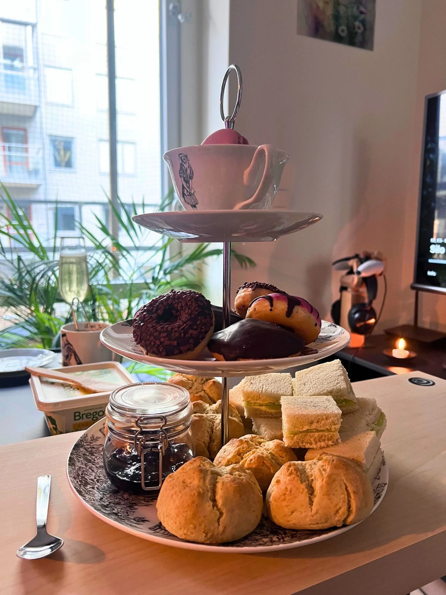 A three-tiered afternoon tea stand on a light wooden table. The top tier has a white teacup with a black illustration on a saucer containing a pink macaron.