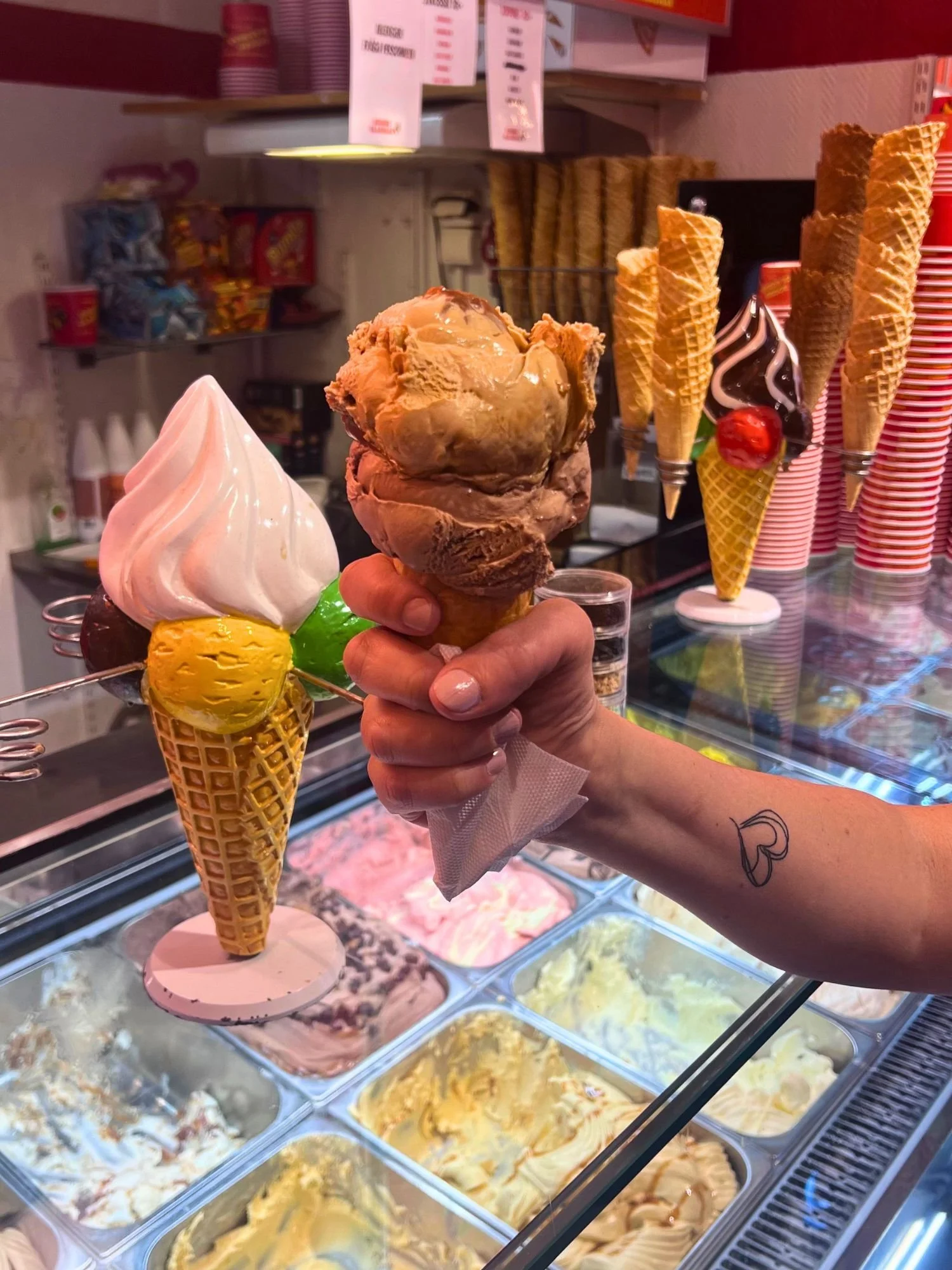 A hand holding a waffle cone with two large scoops of ice cream. The bottom scoop is dark chocolate, and the top scoop is a light brown coffee caramel flavor with a visible swirl.
