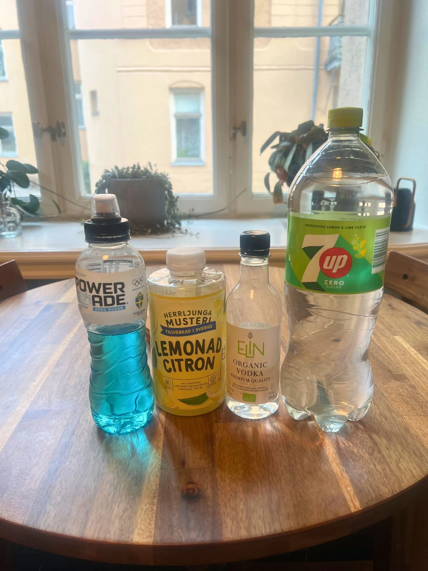 Ingredients for a blue cocktail on a wooden table, including blue Powerade, lemonade, vodka, and lemon-lime soda, with a window and plants in the background.