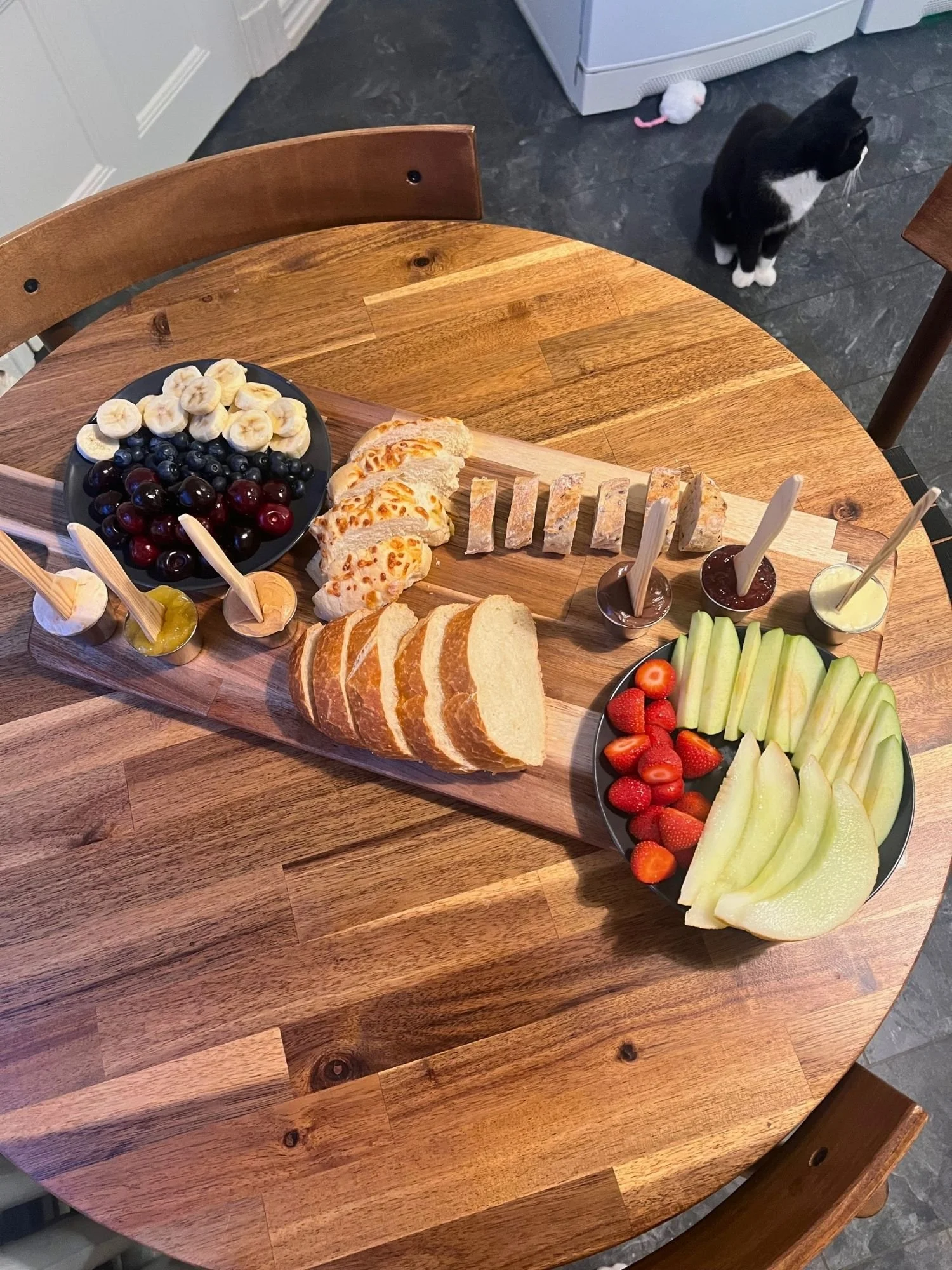 High-angle shot of a long wooden serving board on a round wooden table, arranged with sliced bread and plates of fresh fruit.