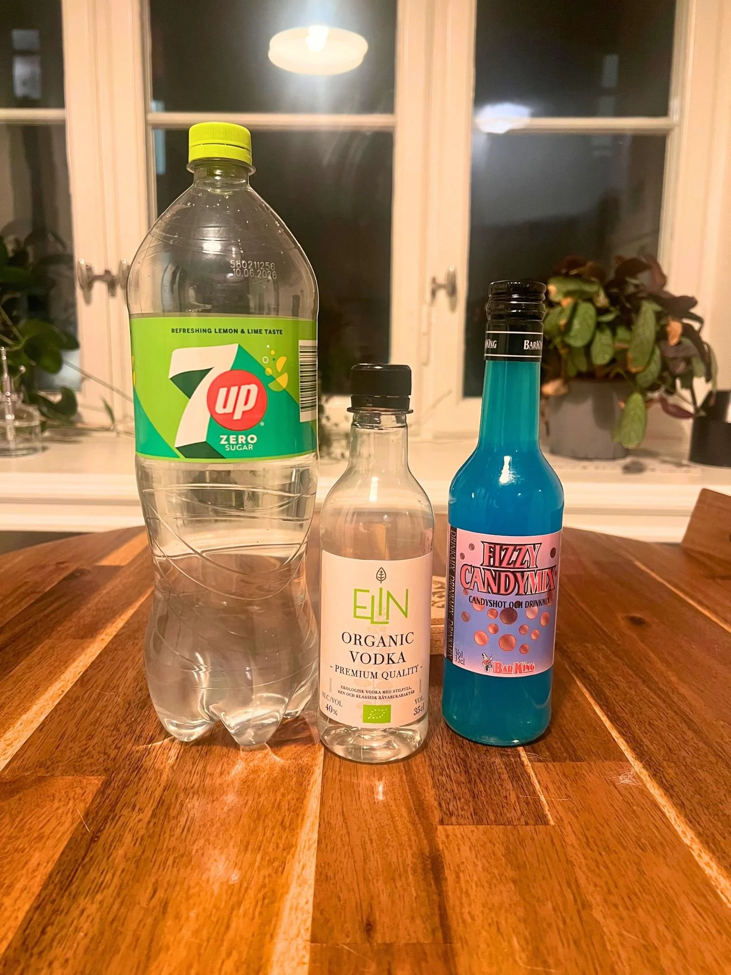 Ingredients for a blue cocktail on a wooden table, including lemon-lime soda, vodka, and blue Fizzy Candymix mixer, with a window and plants in the background.
