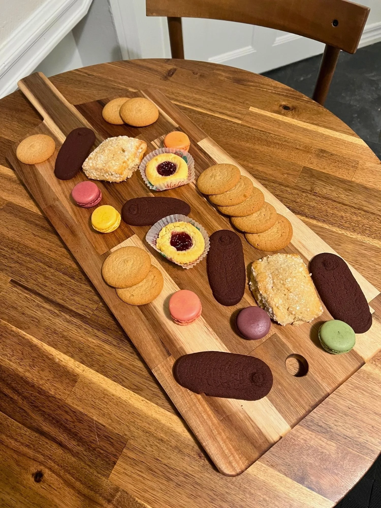 A long wooden serving board with a handle lies diagonally across a round wooden table, neatly displayed with an array of cookies.