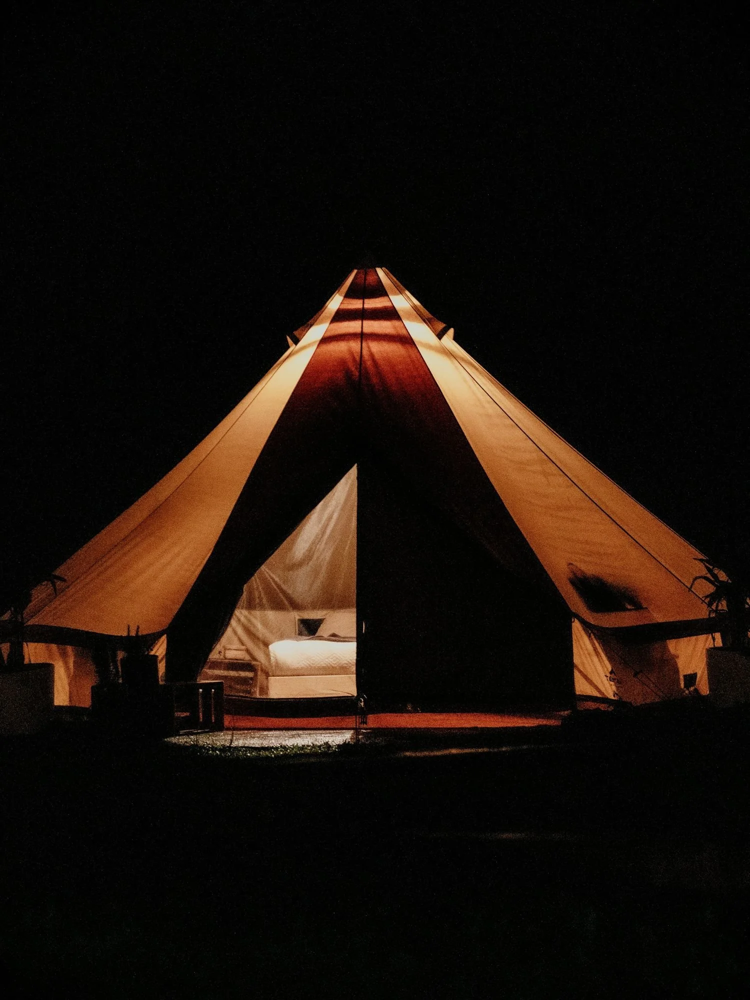 A large canvas bell tent glows with warm, golden light against a completely black night background. The front flaps of the tent are partially drawn back, revealing a glimpse of a neatly made bed with white linens inside the illuminated interior.