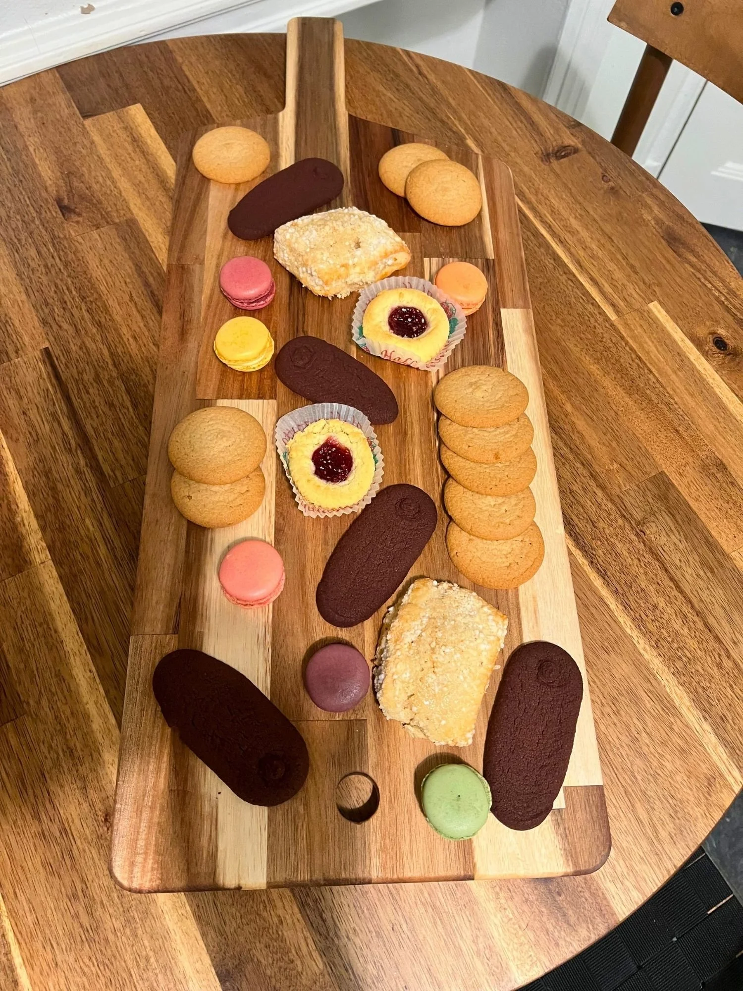 A rectangular wooden serving board lies vertically across a round wooden table, covered with an assortment of cookies, pastries, and colorful macarons.