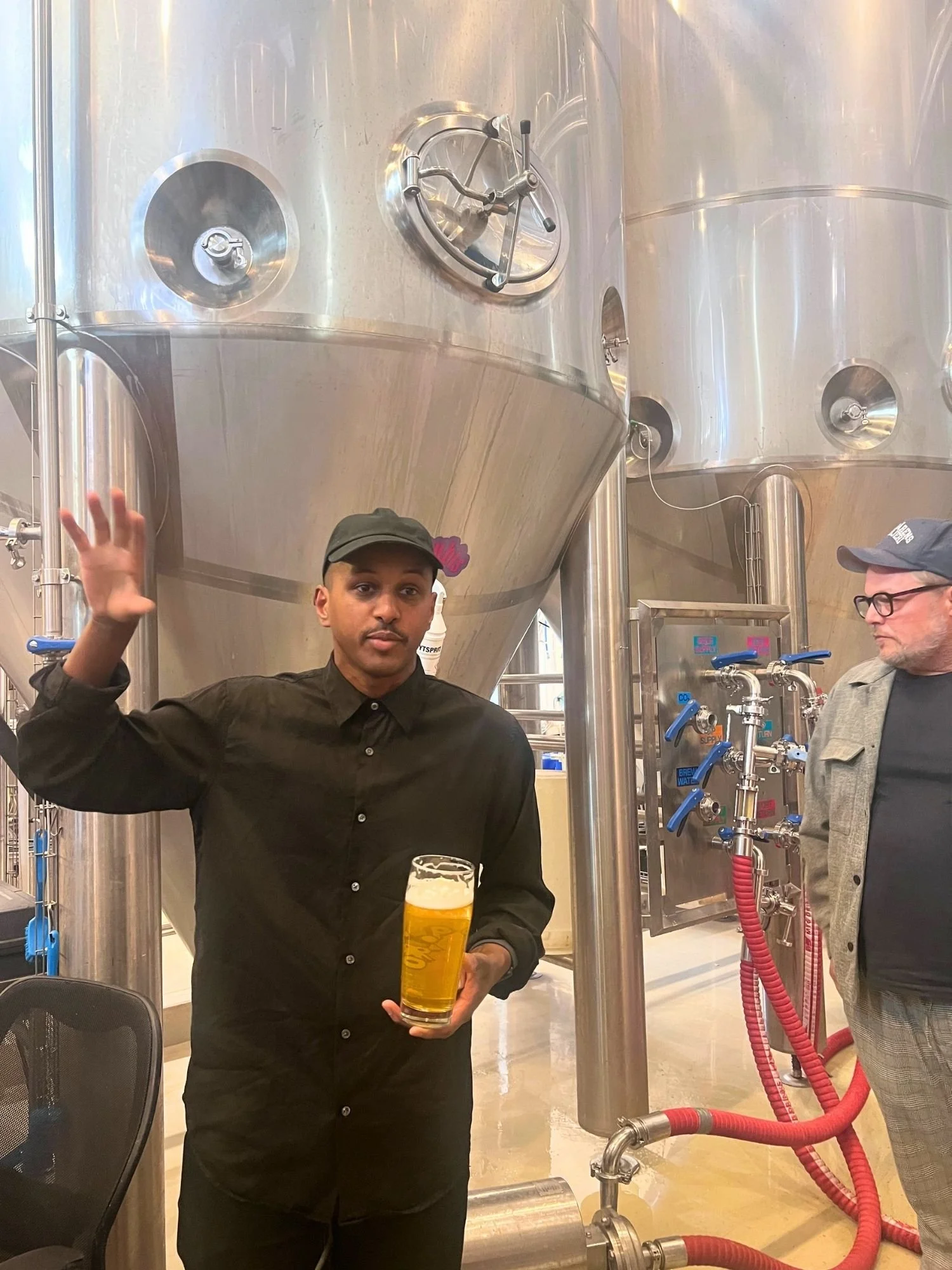 A man wearing a black long-sleeve button-up shirt and a black baseball cap stands in a brewery, gesturing with his raised right hand while holding a glass of draft beer in his left.