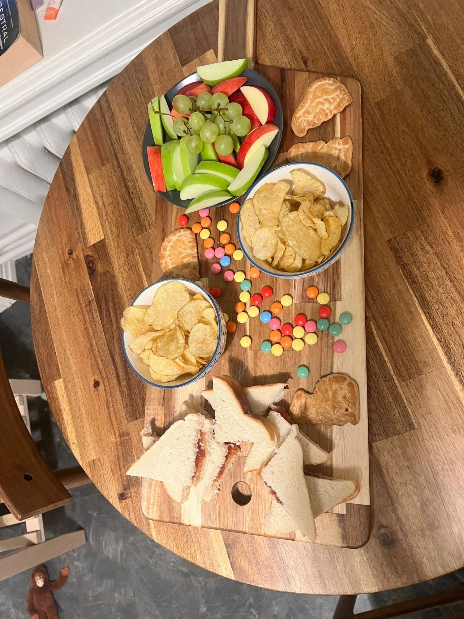 A kid-friendly vegetarian charcuterie board arranged on a wooden serving board, featuring sliced apple, green grapes, potato chips, colorful candy, jam sandwiches cut into triangles, and dinosaur cookies.