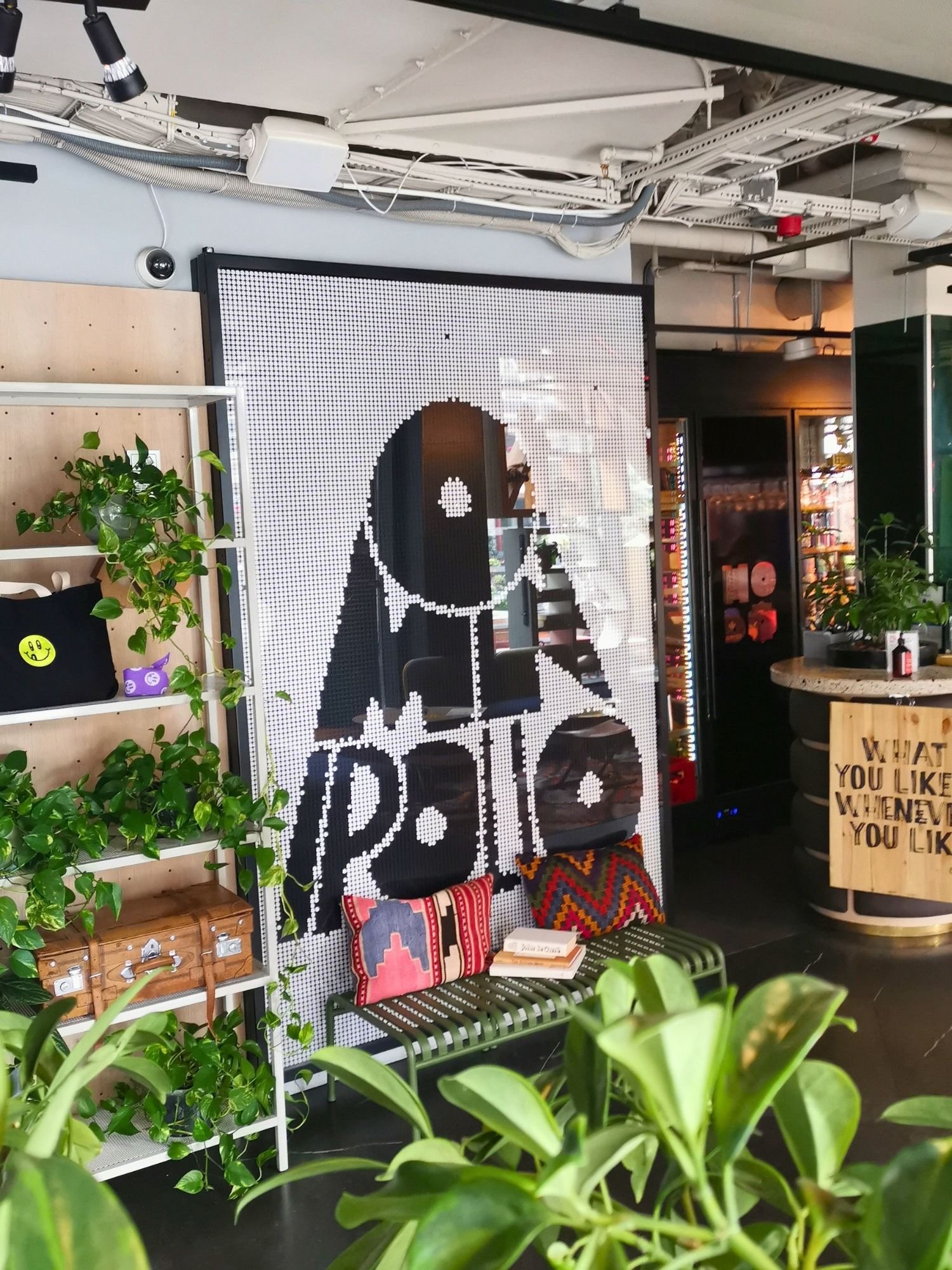 A large, black-and-white pixelated mural spelling "OMNIPOLLO" displayed on a feature wall inside a modern, plant-filled venue. Below the wall sits a green metal bench adorned with colorful patterned pillows and a small stack of books.