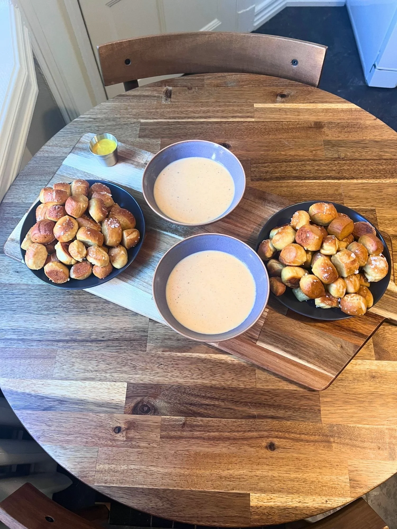 High-angle, overhead view of a wooden serving board set on a round wooden table, holding two plates of salted pretzel bites, two bowls of cheese sauce, and a small cup of mustard sauce.