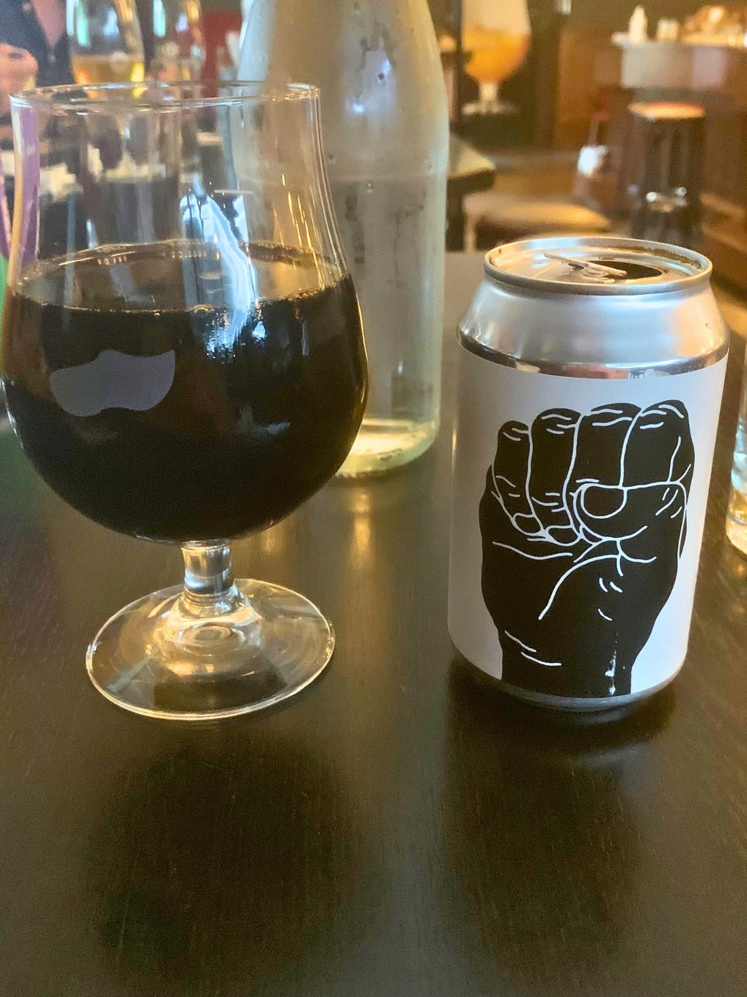 A dark beer in a stemmed glass next to a can featuring a bold black-and-white fist design in a restaurant with other drinks in the background.