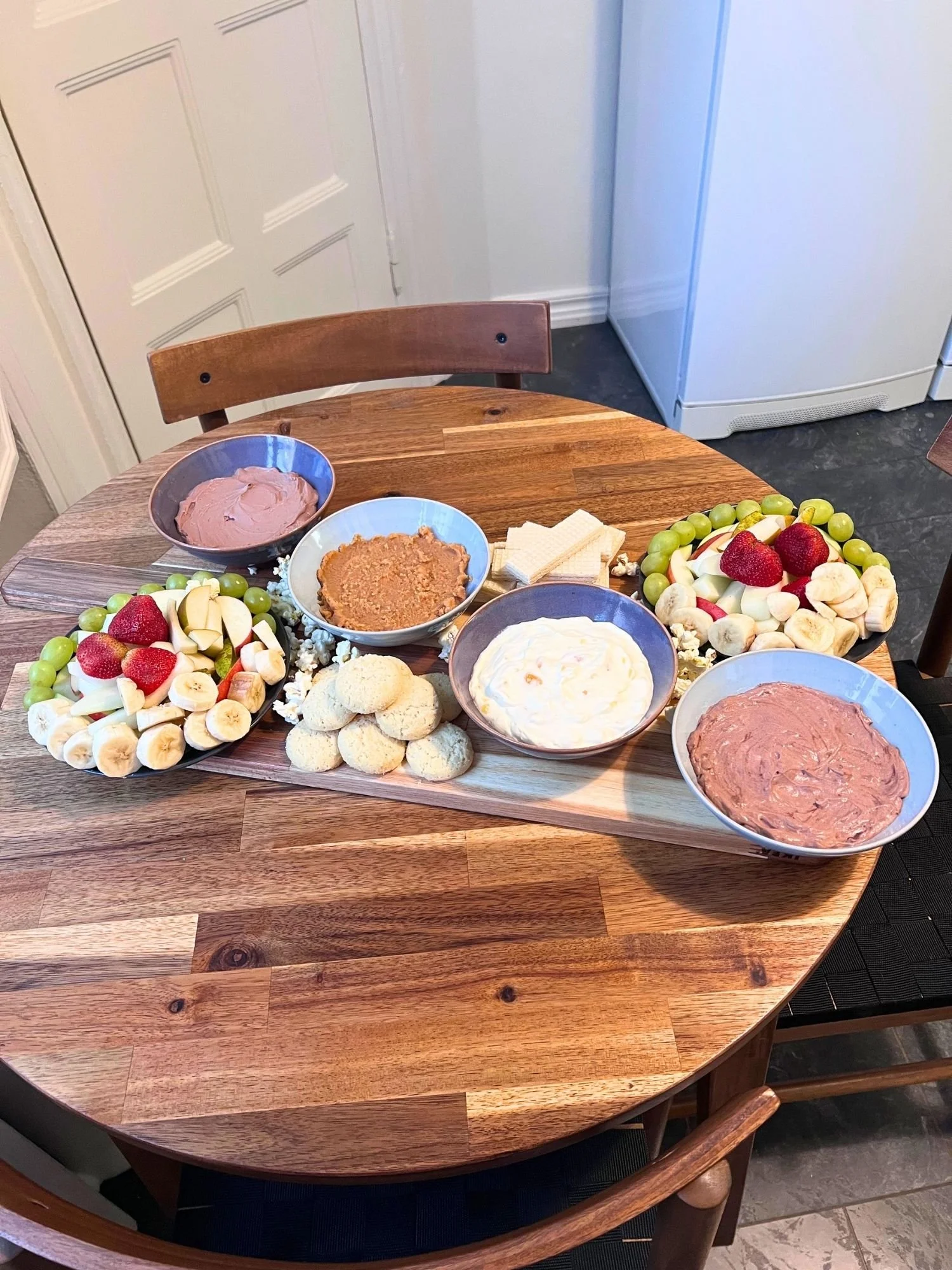 A wooden table set with a serving board holding two plates of sliced fresh fruit, four ceramic bowls of sweet dessert dips, cookies, and wafer biscuits.