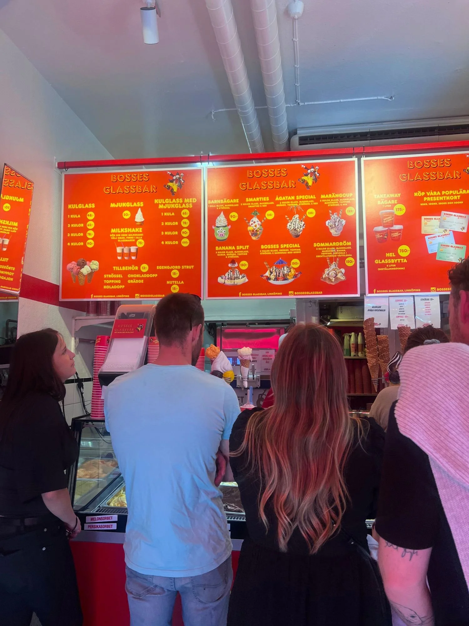 A queue of customers looking up at a large, vibrant red menu board inside an ice cream shop.