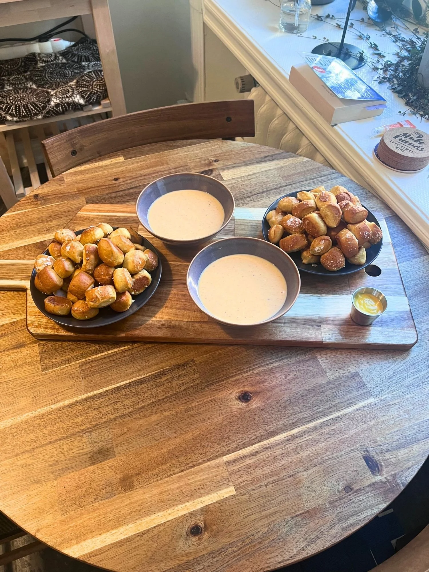 A wooden table set with a serving paddle holding two plates of warm pretzel bites, two bowls of warm cheese sauce, and a small cup of yellow mustard.