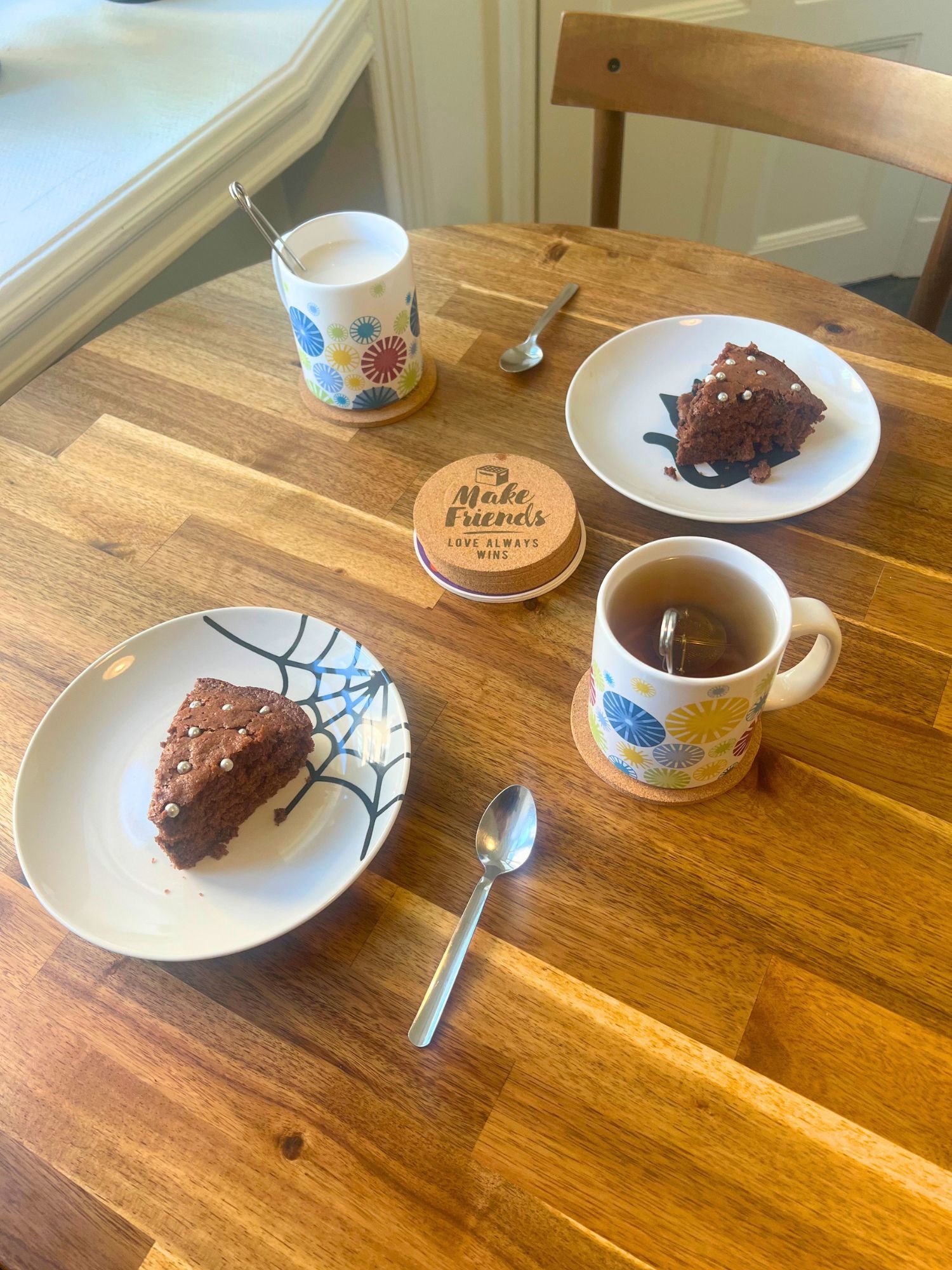Two slices of chocolate sockerkaka topped with round, silver cake decorations served on small plates, alongside mugs of tea and spoons on a wooden table, set for a cozy fika.