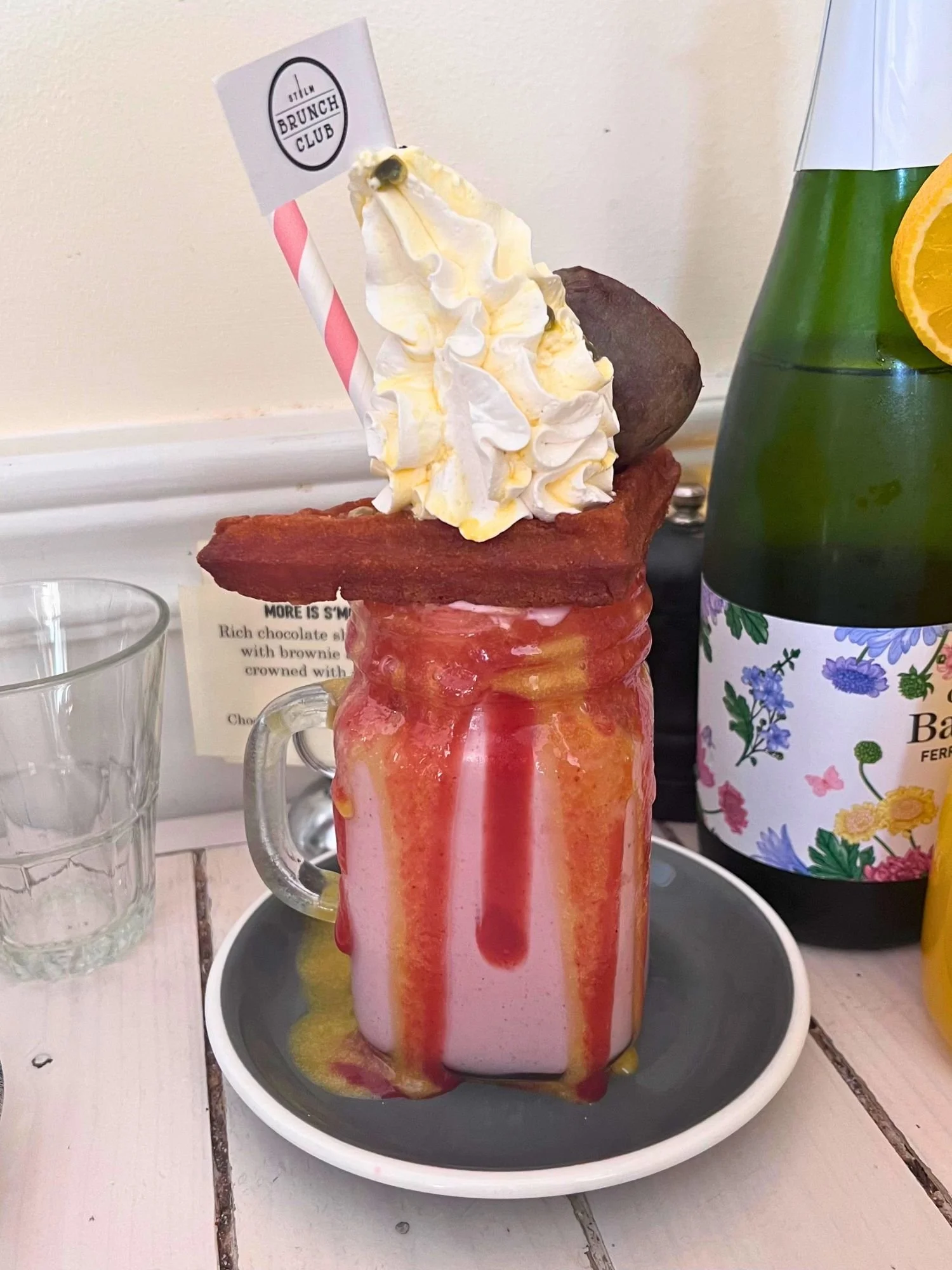 An over-the-top strawberry freakshake served in a glass mason jar on a grey saucer, overflowing with vibrant red strawberry and yellow passion fruit syrups dripping down the sides.