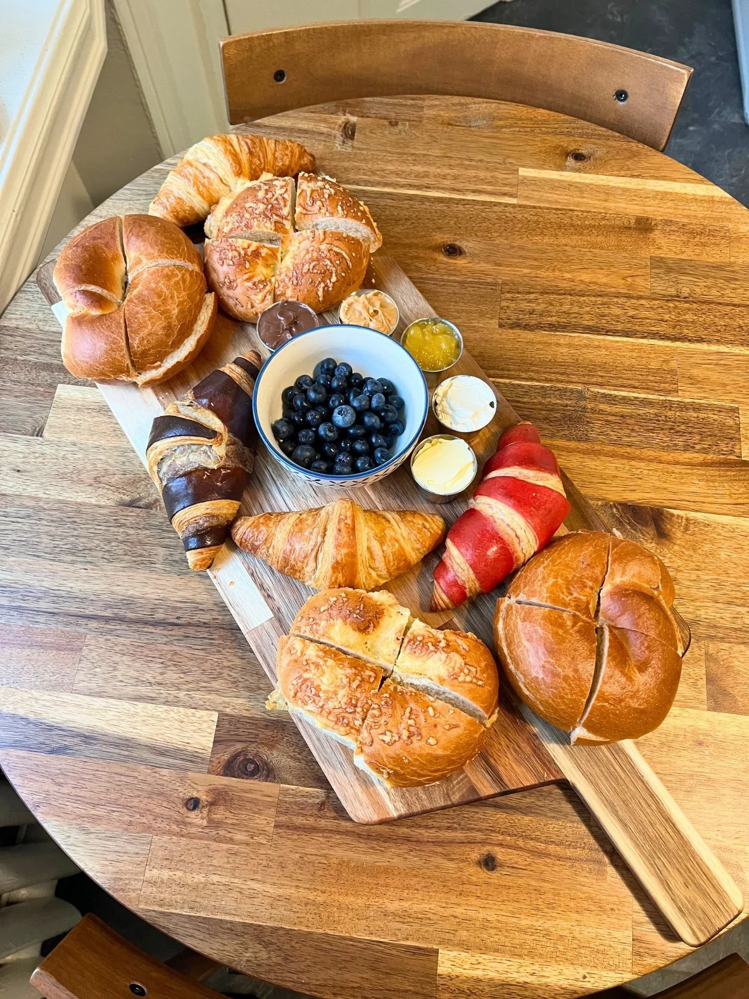 High-angle shot of a long wooden serving board on a round wooden table, arranged with a variety of croissants and bagels, a small bowl of fresh blueberries, and small metal ramekins with butter and spreads.