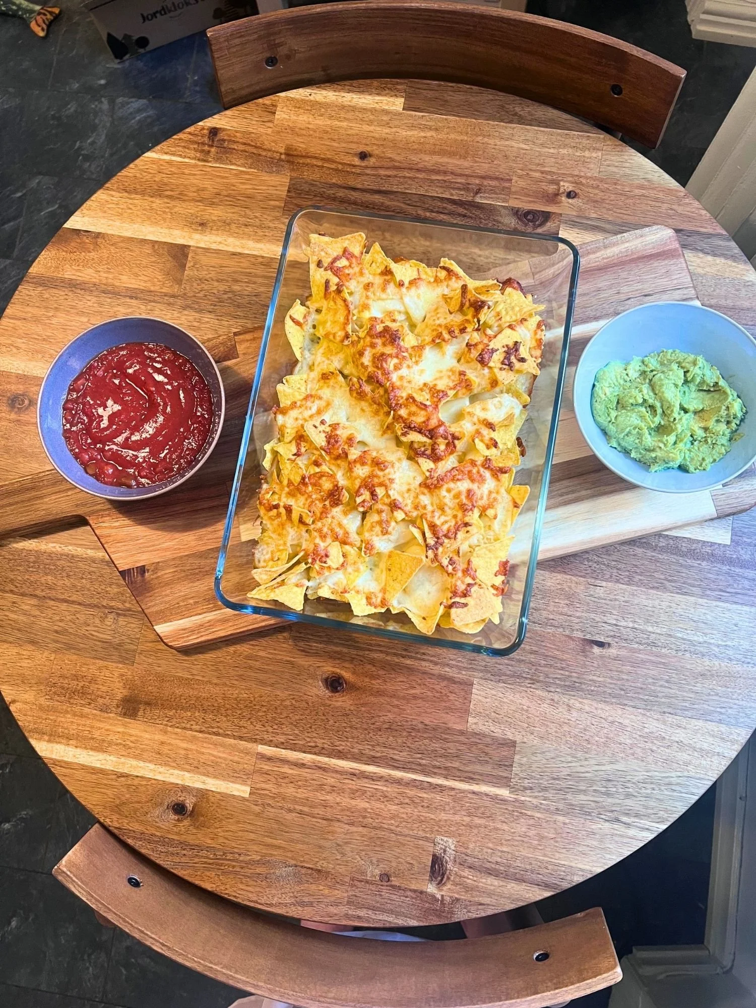 Direct overhead view of a glass baking dish with baked cheese nachos, accompanied by bowls of salsa and guacamole on a wooden serving board.