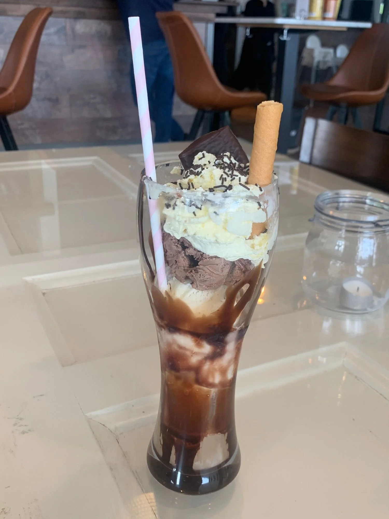 A tall ice cream sundae in a glass on a white table, layered with chocolate syrup, vanilla and chocolate ice cream, and whipped cream.
